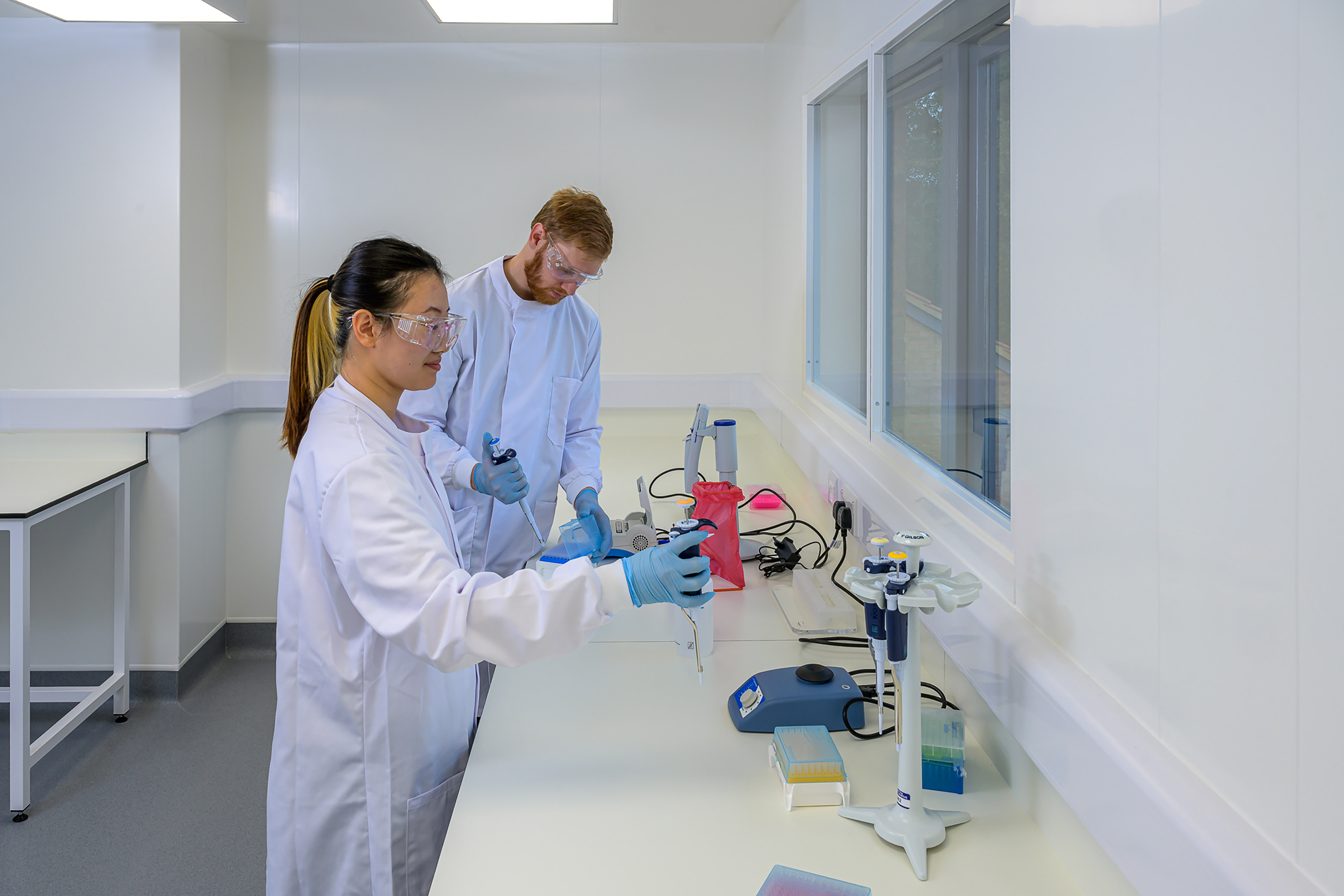 Two scientists in lab coats and safety goggles use pipettes at a lab bench, surrounded by various laboratory equipment and supplies, in a bright, sterile laboratory environment.