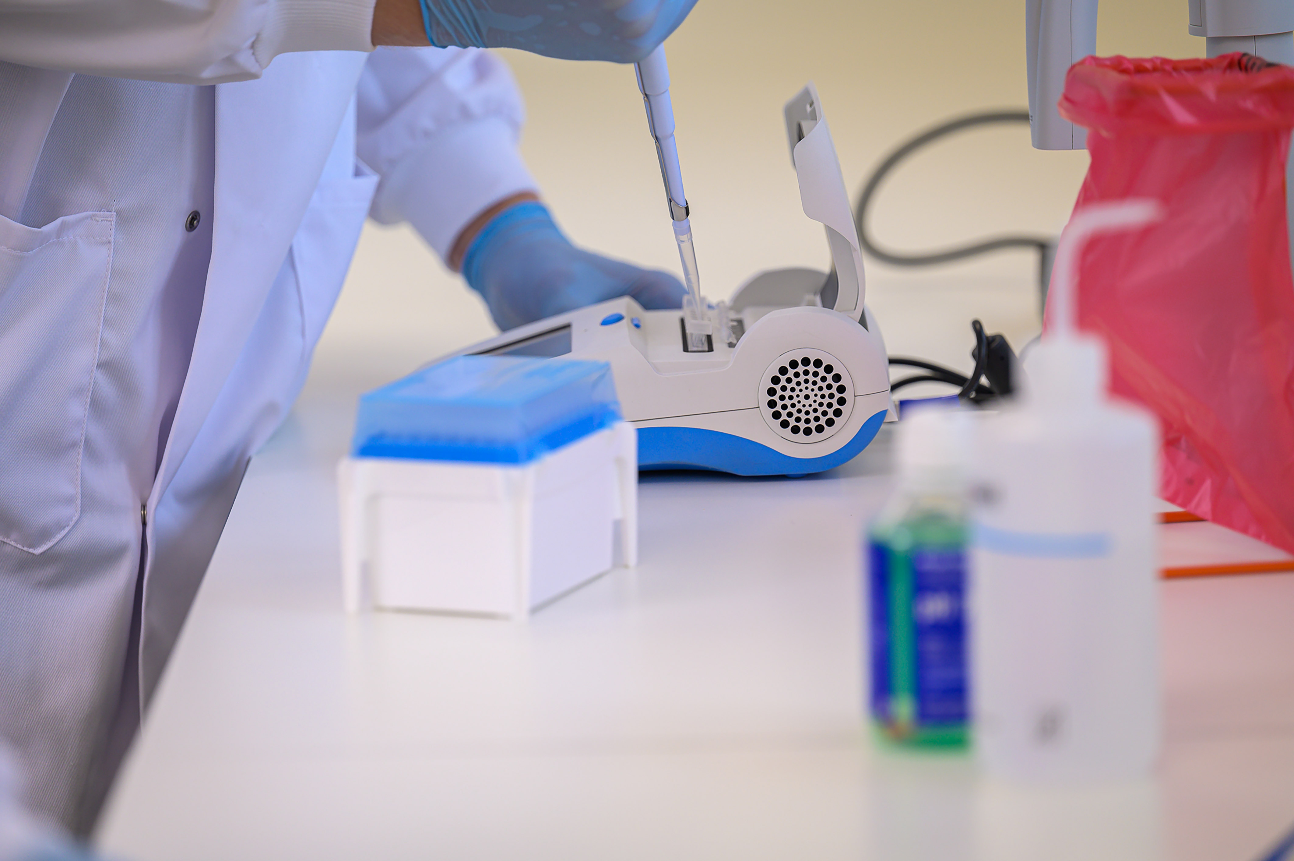 A laboratory device on a table, being operated with a pipette by a gloved hand, surrounded by containers, a computer, and a red bag in a research setting.