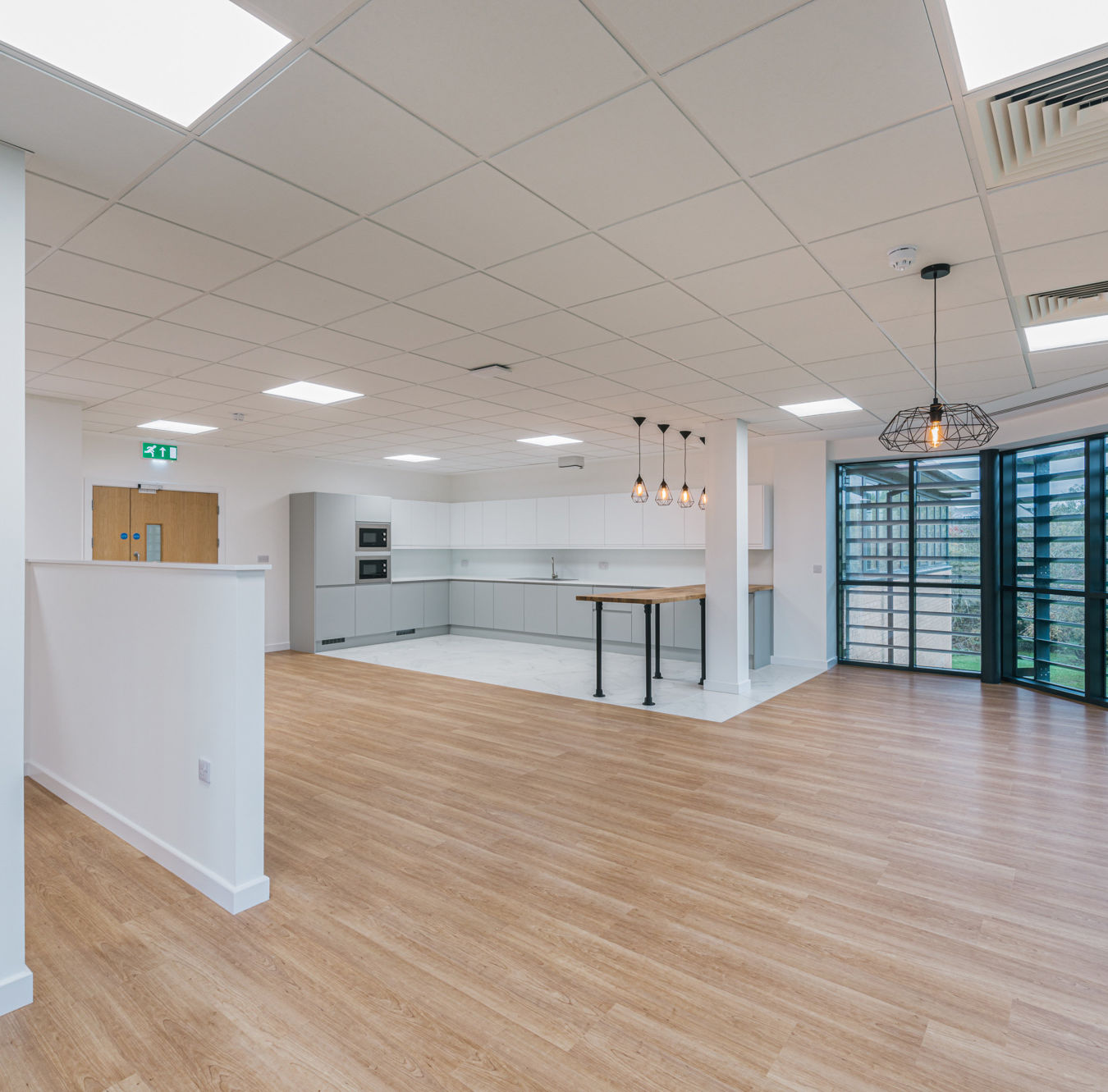 Spacious modern kitchen features white cabinets, a wooden island, and pendant lights, set in a well-lit room with wood flooring and large windows overlooking greenery.