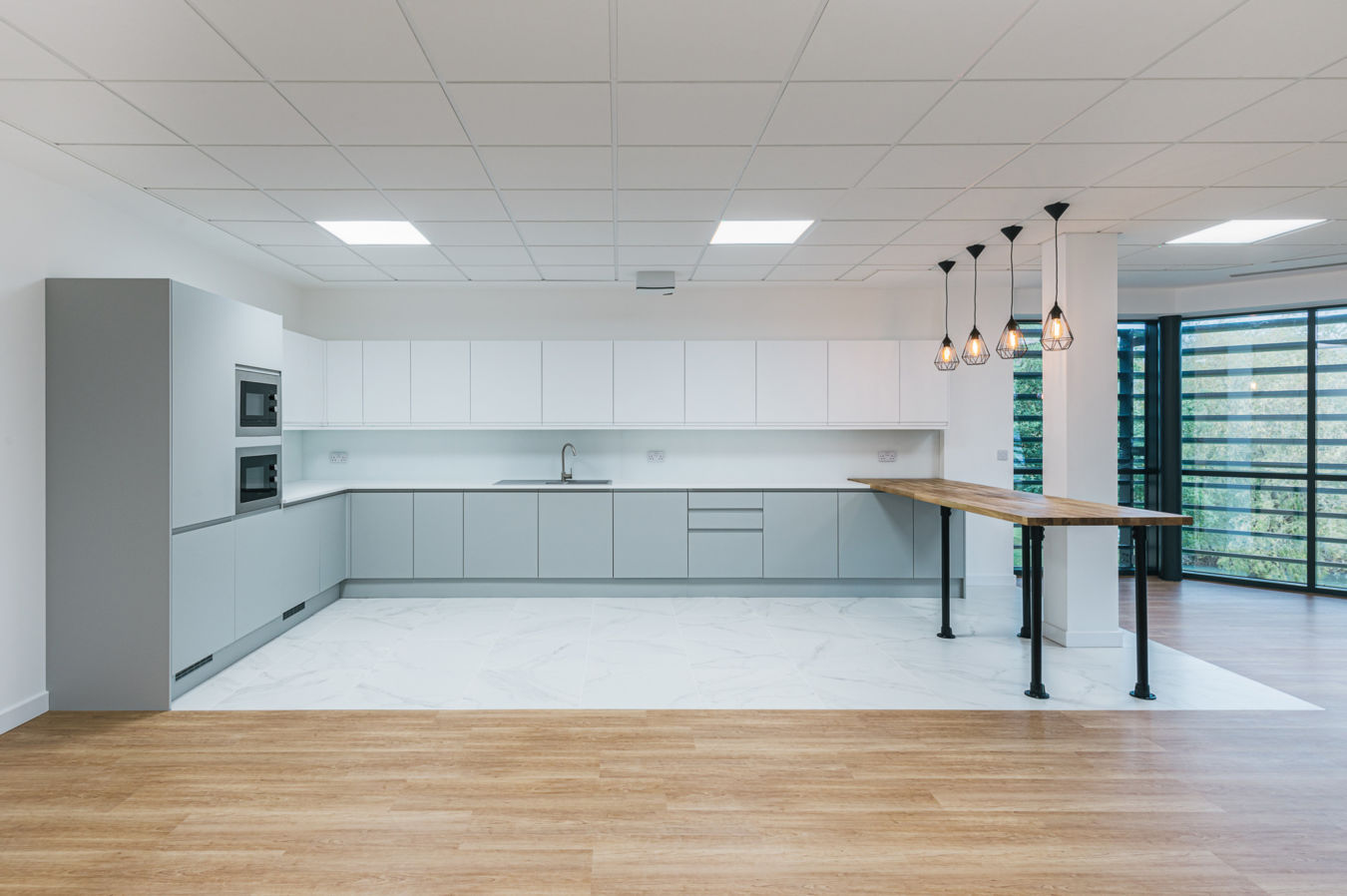 Modern kitchen with light gray cabinetry, under-counter oven, and a stainless steel sink. Wooden counter extends with pendant lights above, while glass windows illuminate the space.