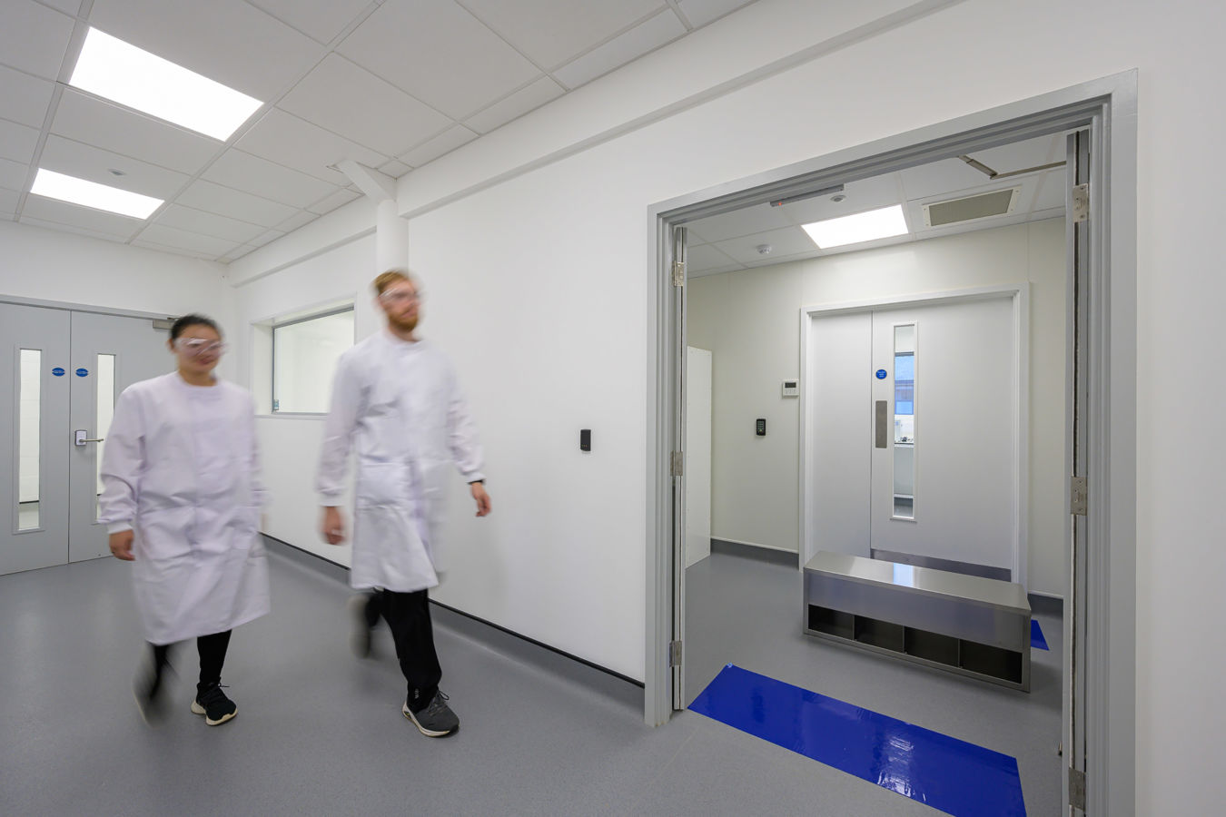 Two people wearing white lab coats and safety goggles walk through a clean, bright corridor with white walls and gray floors, leading to an industrial metal door.