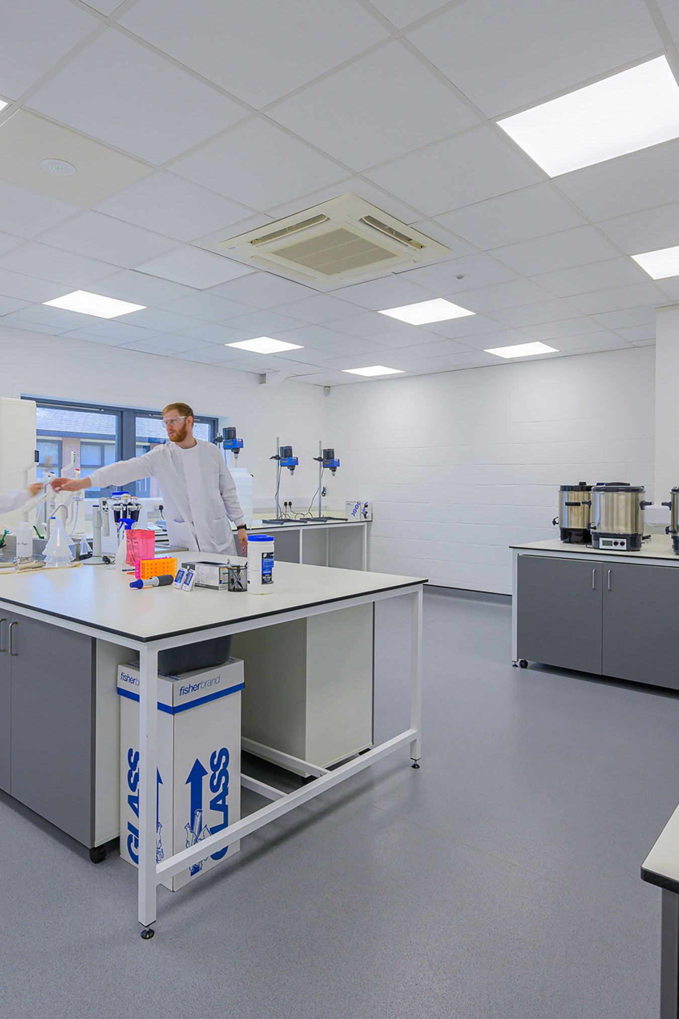 A person wearing a lab coat works in a modern laboratory setting, with various scientific equipment on the tables. Text on a box reads "fisherbrand GLASS."