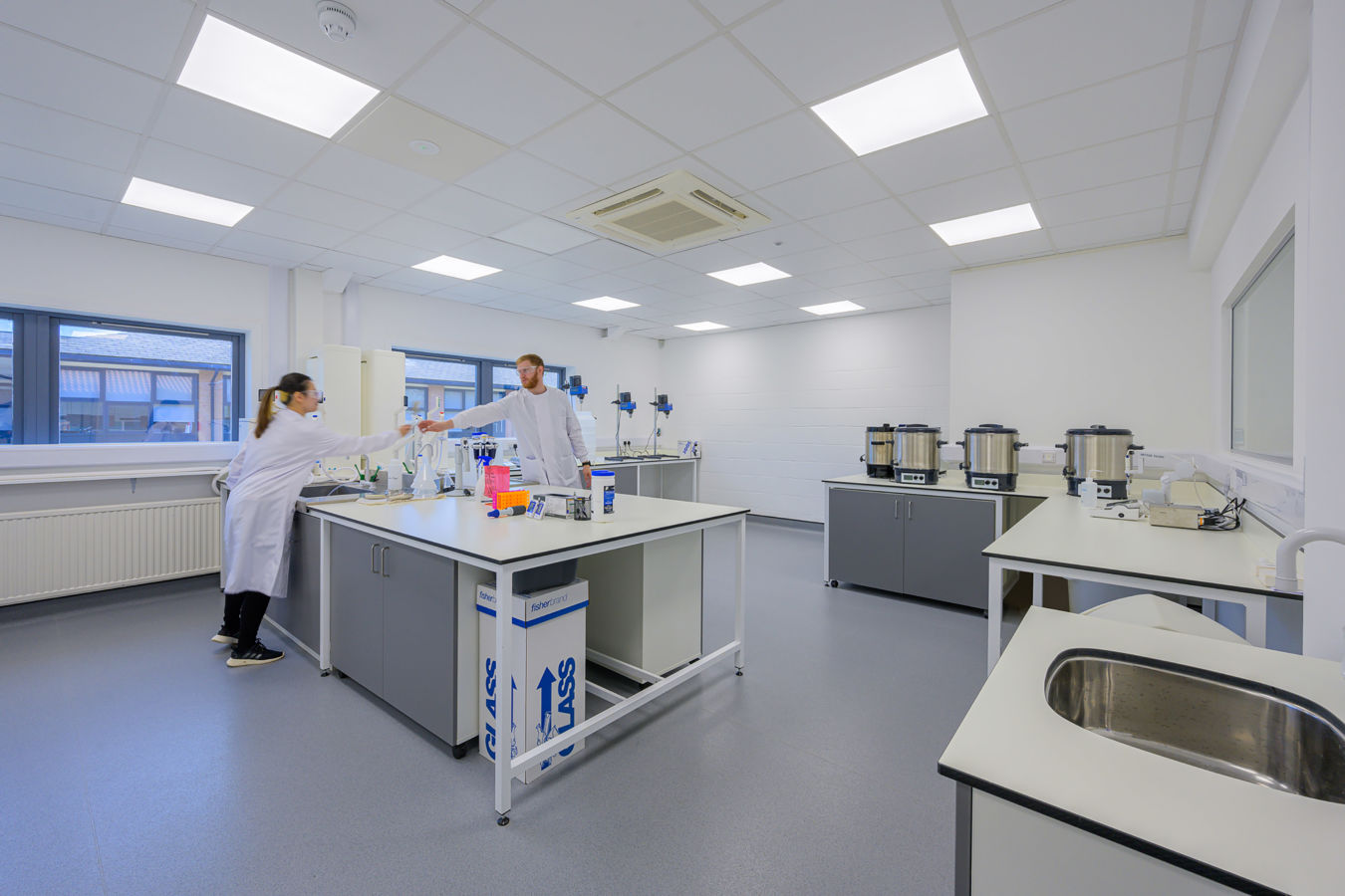 A person wearing a lab coat works in a modern laboratory setting, with various scientific equipment on the tables. Text on a box reads "fisherbrand GLASS."