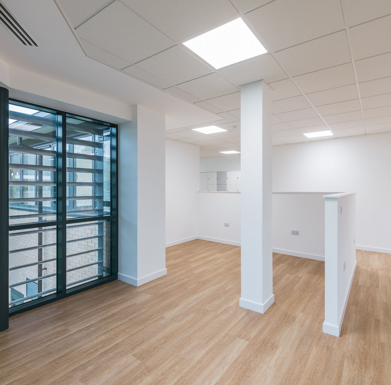 A modern, empty room with wooden flooring features a large window with black frames and a white ceiling with recessed lighting, creating a bright, open office space.