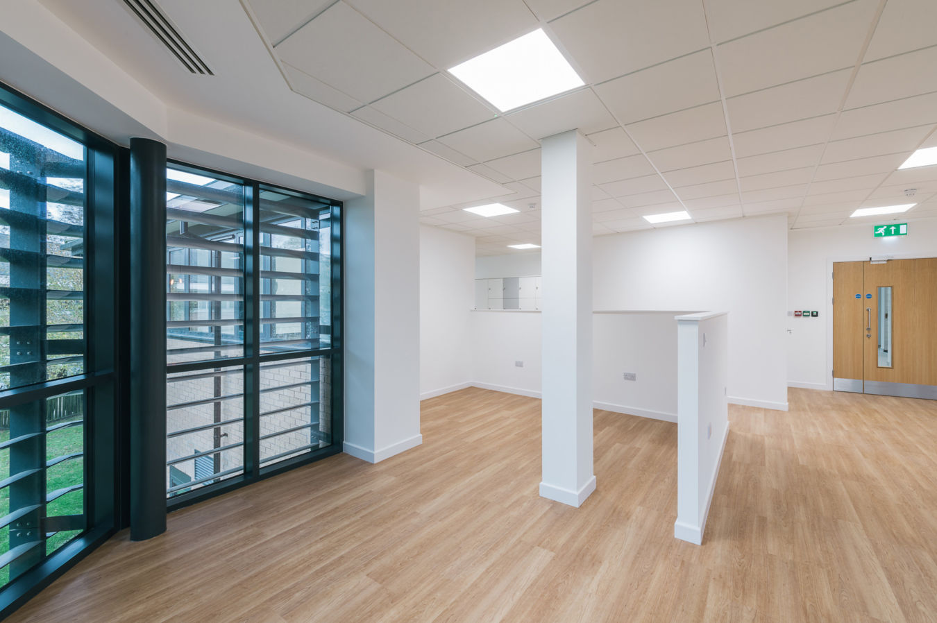 A modern, empty room with wooden flooring features a large window with black frames and a white ceiling with recessed lighting, creating a bright, open office space.