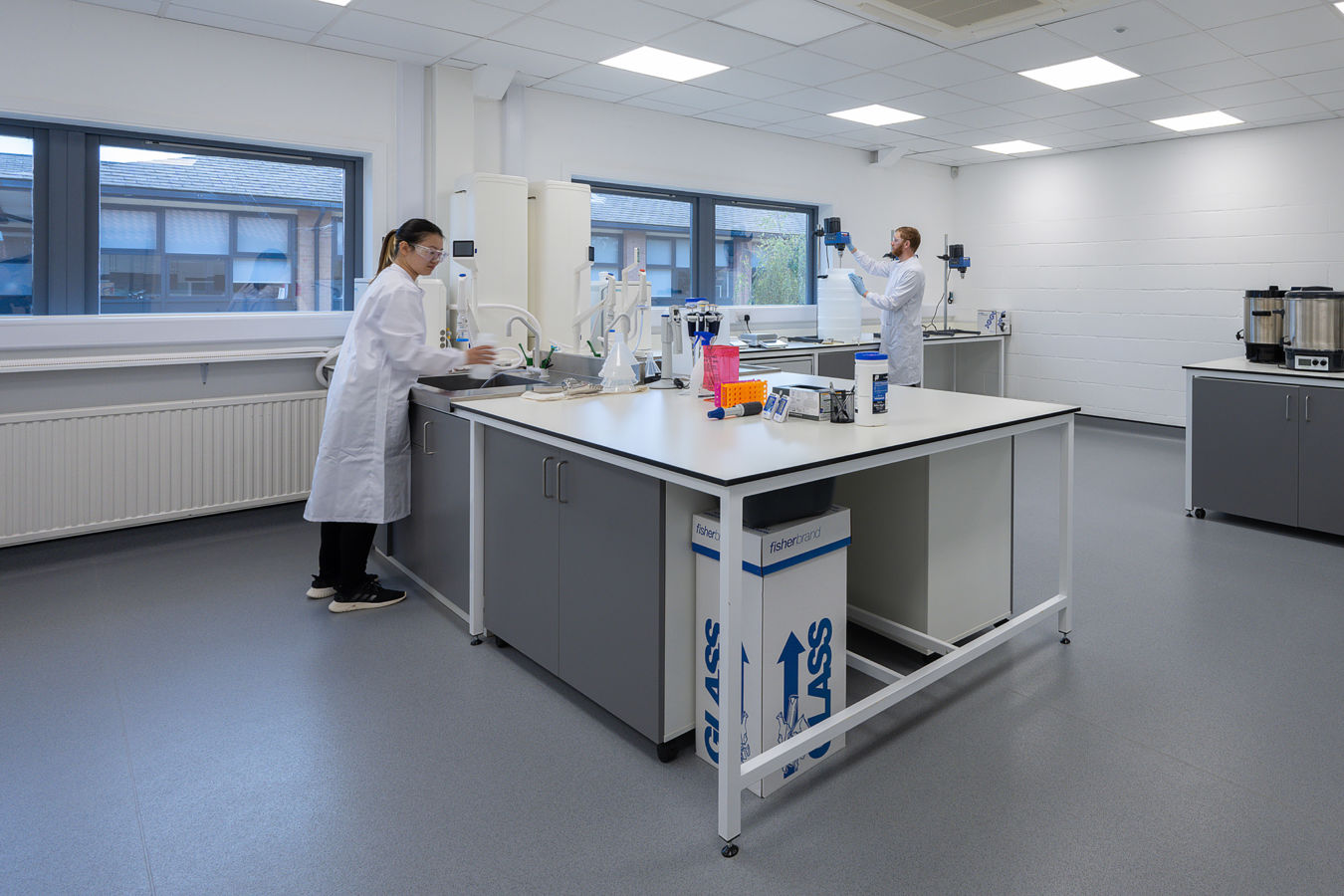 Laboratory with two scientists in white lab coats working at a central workstation equipped with lab equipment, under bright lighting and surrounded by large windows.