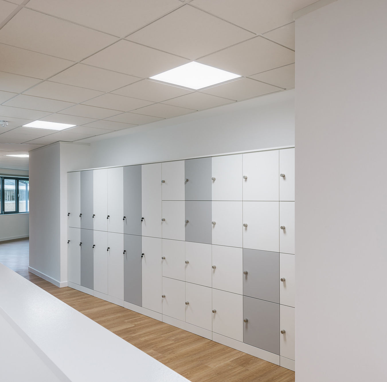 brightly lit corridor with white and gray lockers lining the wall. The floor is light wood, and there's a large window in the background, creating an open, clean, and modern atmosphere