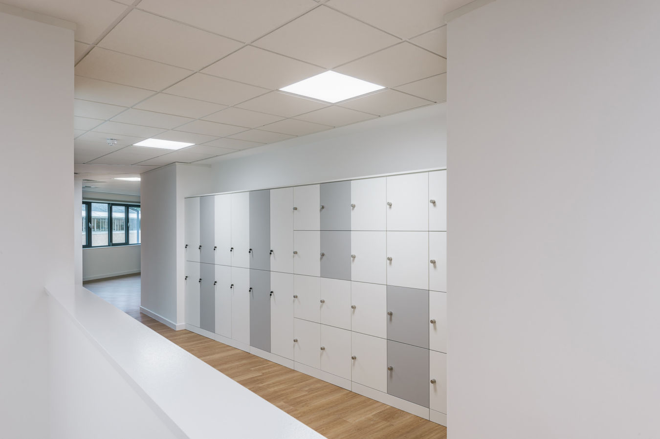 brightly lit corridor with white and gray lockers lining the wall. The floor is light wood, and there's a large window in the background, creating an open, clean, and modern atmosphere