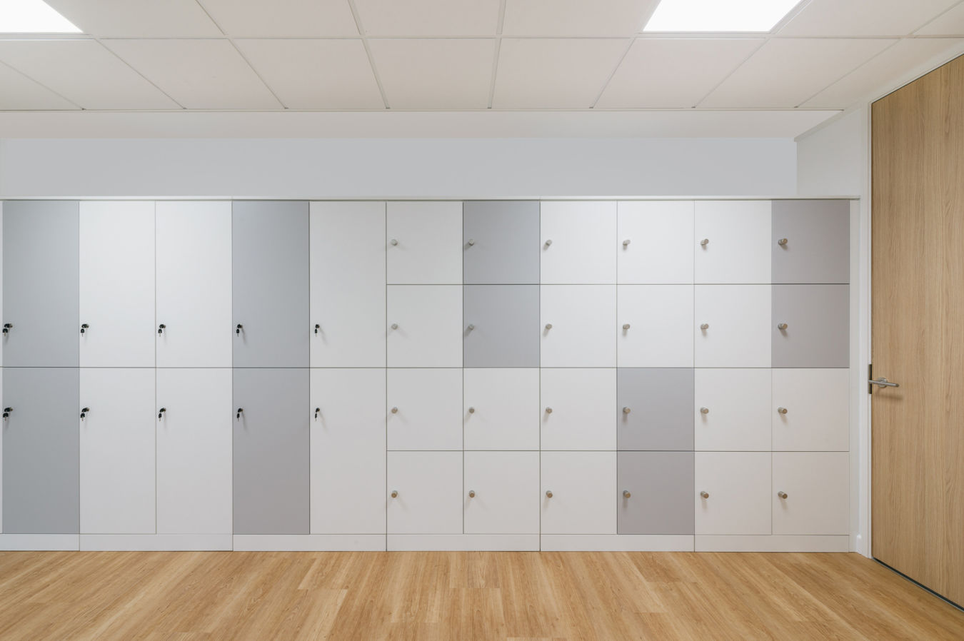 wall of modern, multi-colored lockers in shades of white and gray with wooden handles. The floor is light wood, and a wooden door is on the right