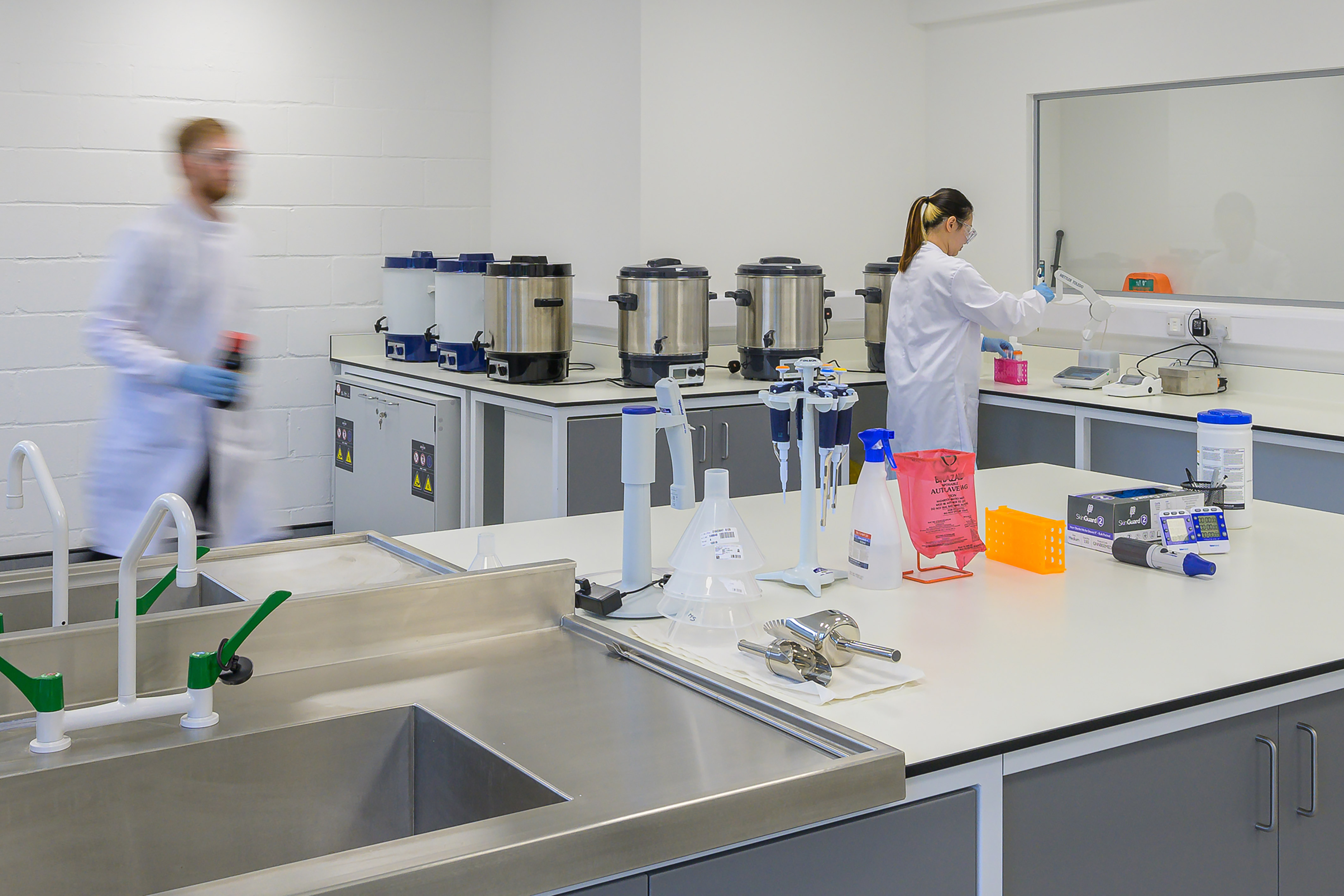 A laboratory with two scientists in white coats, one walking, the other using equipment; surrounded by lab tools, pipettes, cleaning supplies, and stainless steel counters in a bright, modern setting.