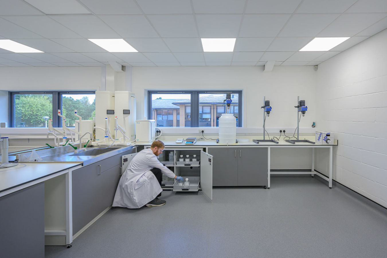 A person in a white lab coat arranges bottles inside a cabinet in a bright, modern laboratory with large windows and scientific equipment on countertops.