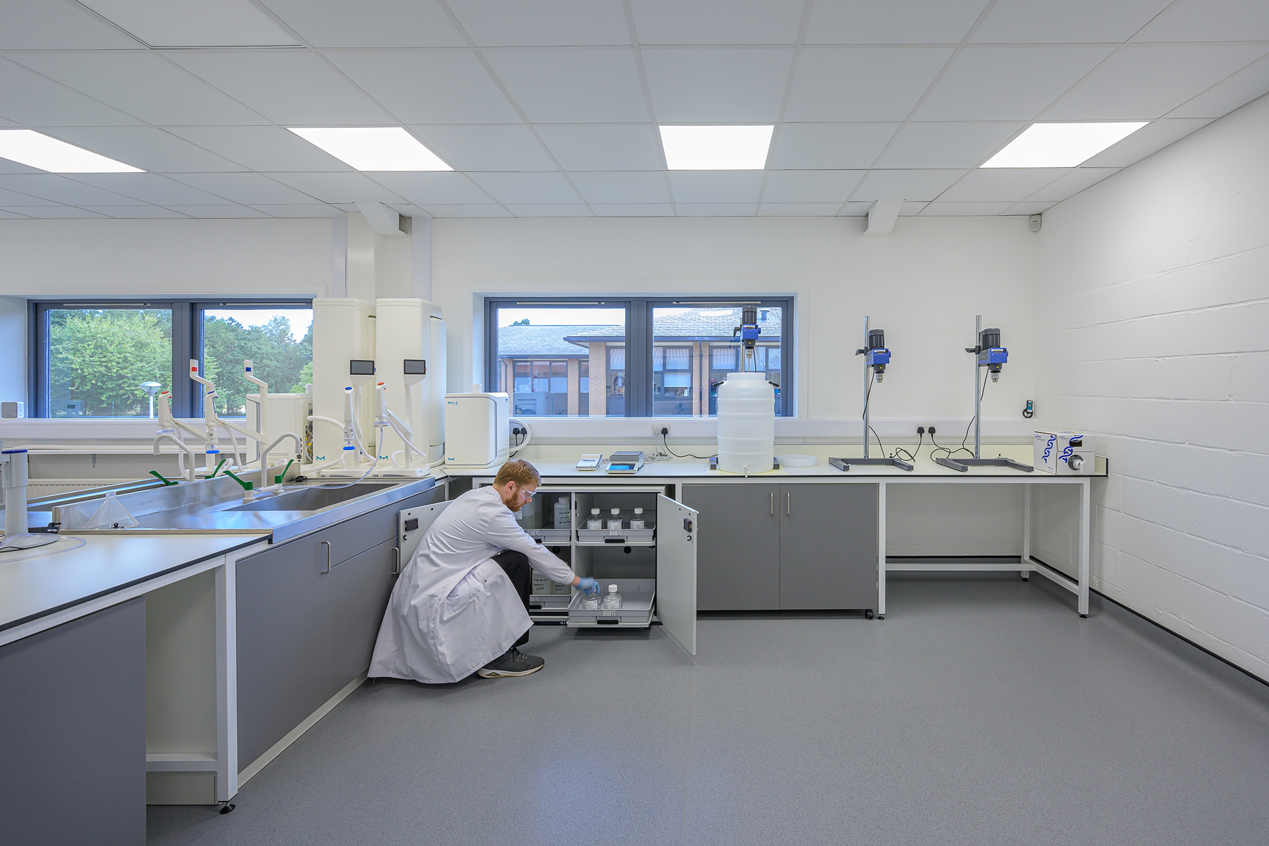 A person in a white lab coat arranges bottles inside a cabinet in a bright, modern laboratory with large windows and scientific equipment on countertops.