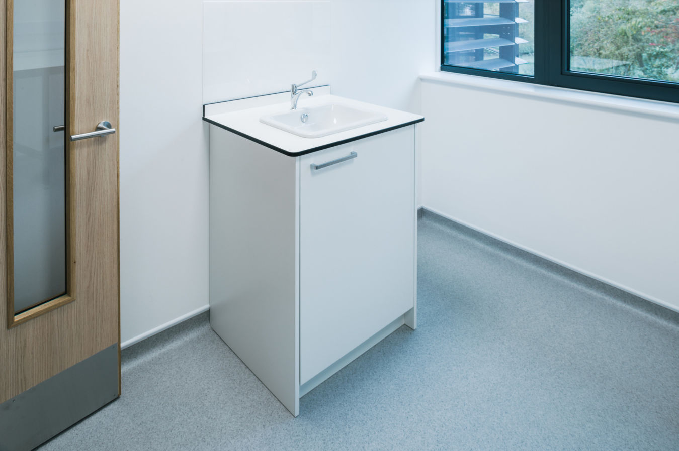 A white sink with a silver faucet is installed on a cabinet in a minimalistic room with grey flooring, a wooden door on the left, and a window on the right.