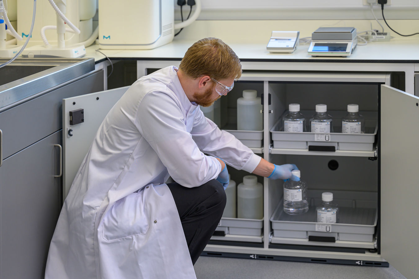 A scientist wearing a lab coat and safety goggles retrieves a bottle from a cabinet in a laboratory, with various bottles and equipment visible on the counter above.