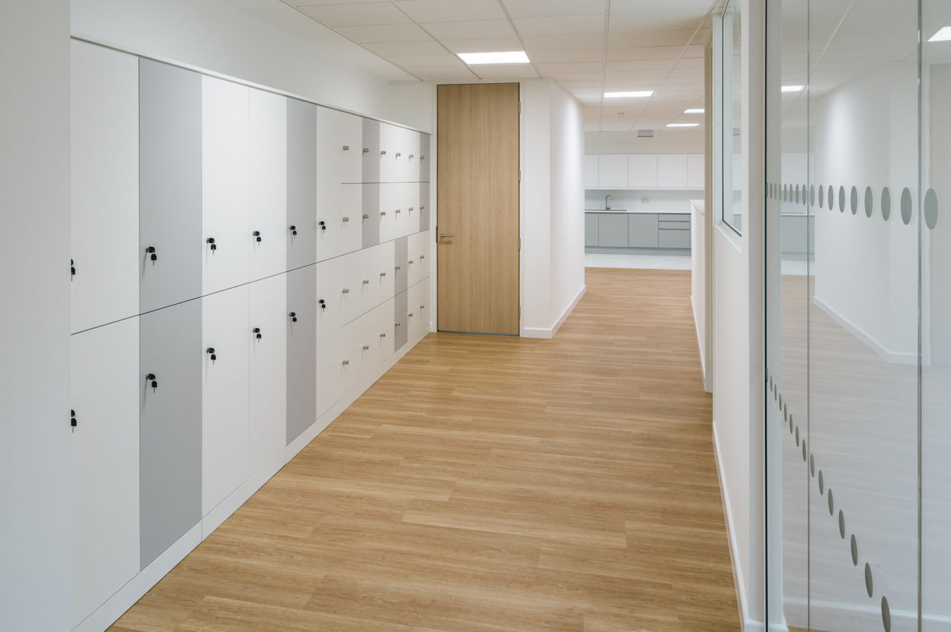 Bright hallway with light wood flooring, white lockers with gray accents on the left, a wooden door at the end, and glass walls on the right. Modern and tidy ambiance
