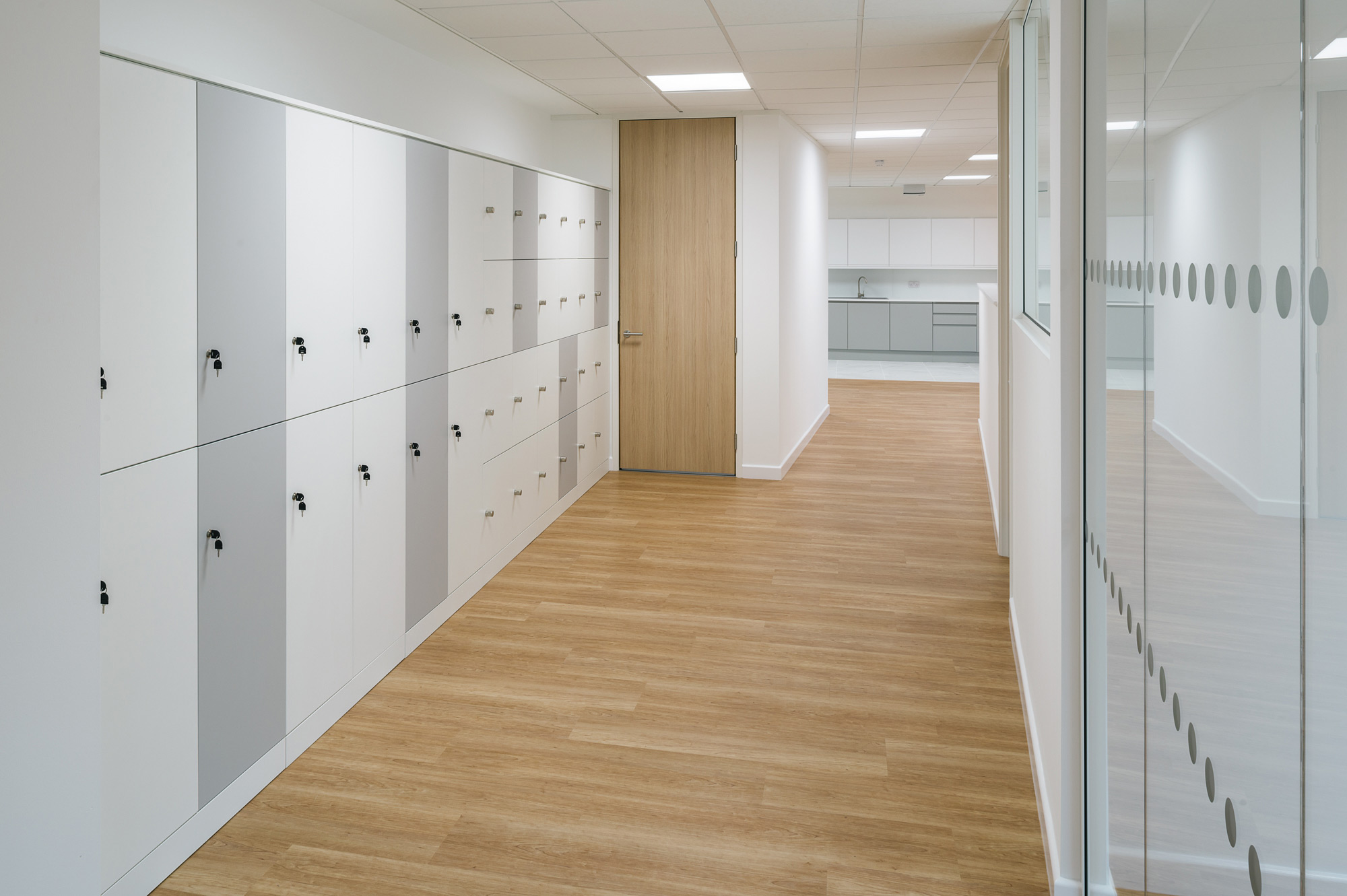 Bright hallway with light wood flooring, white lockers with gray accents on the left, a wooden door at the end, and glass walls on the right. Modern and tidy ambiance