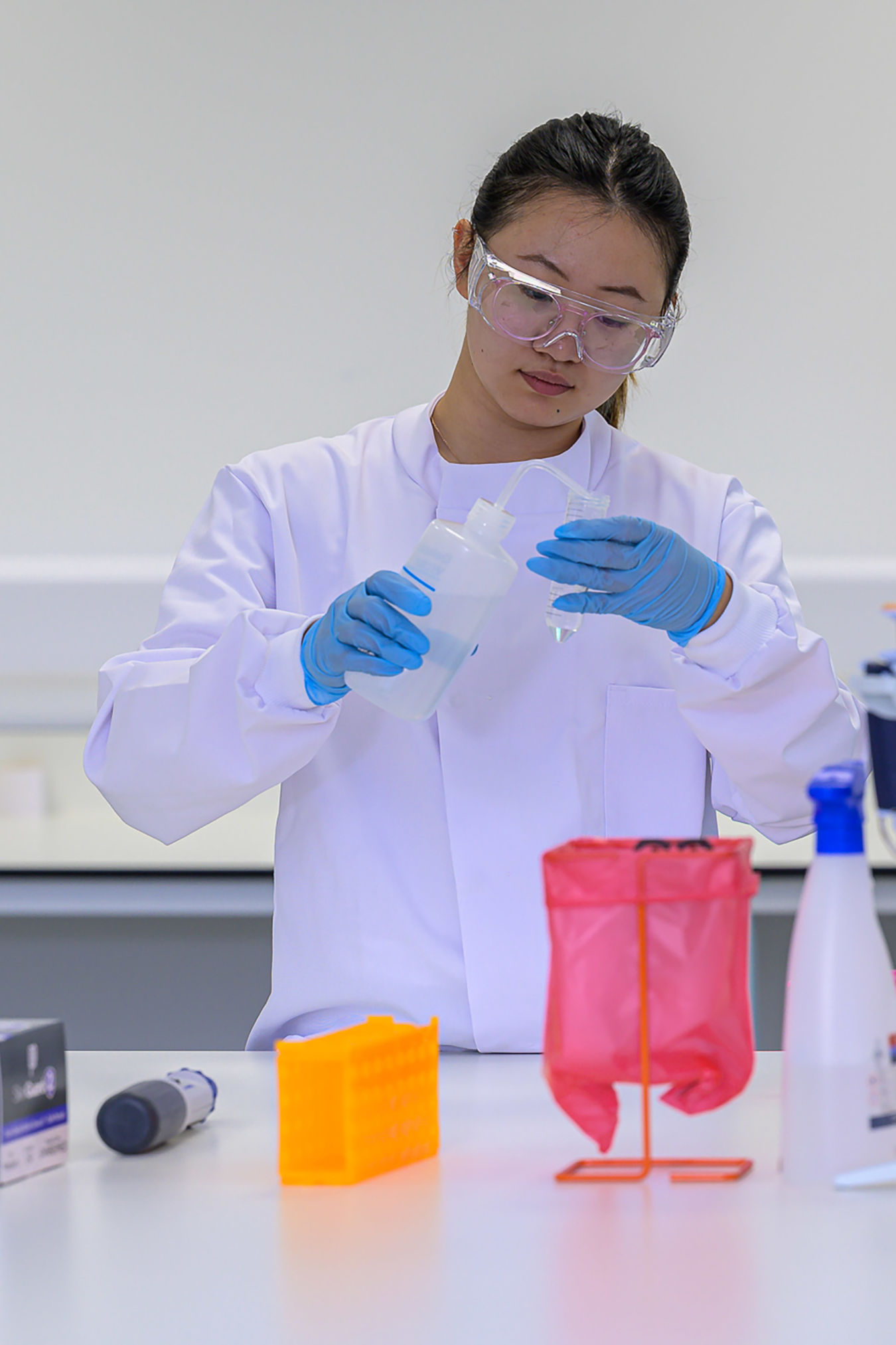 Scientist wearing safety goggles and gloves pours liquid into a container at a lab bench, surrounded by pipettes, a red biohazard bag, and a box labeled "SkinGuard 2".