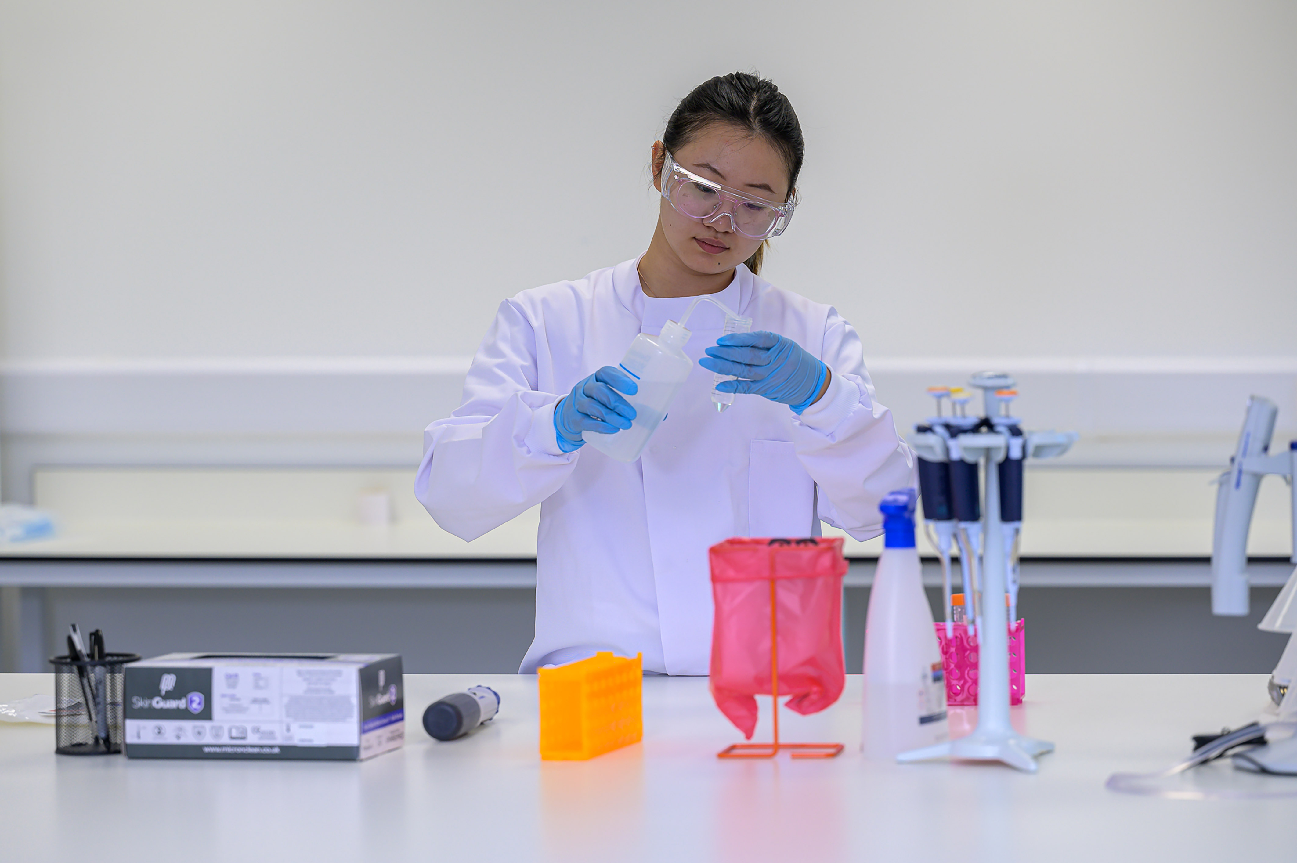 Scientist wearing safety goggles and gloves pours liquid into a container at a lab bench, surrounded by pipettes, a red biohazard bag, and a box labeled "SkinGuard 2".