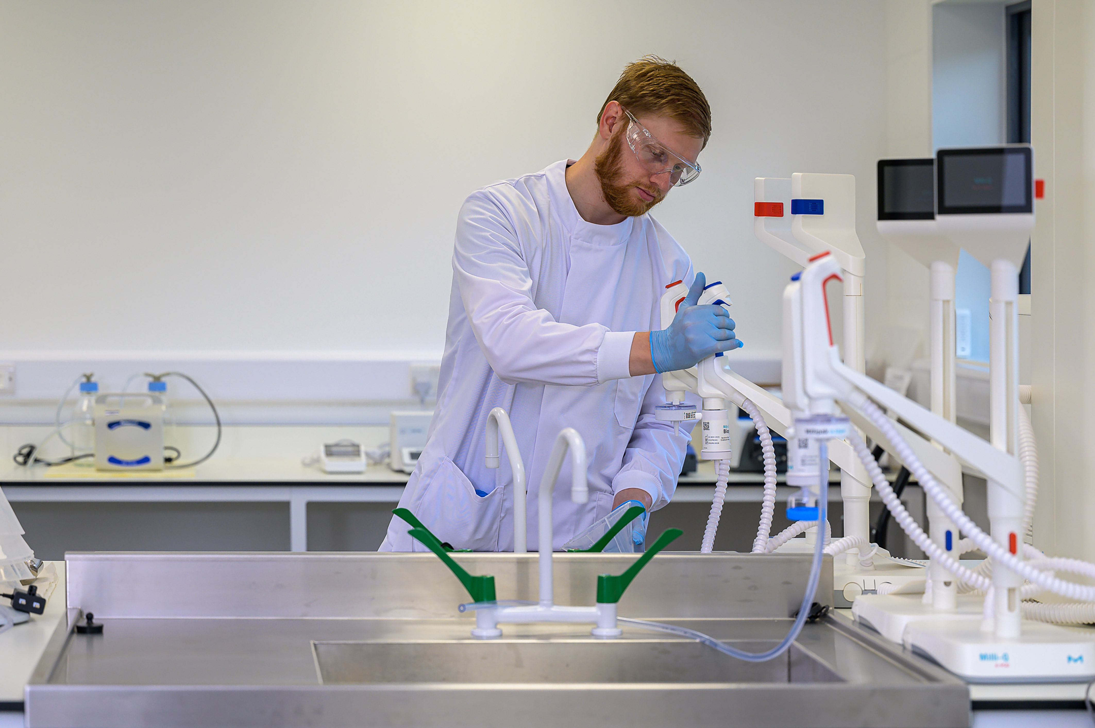 A laboratory technician in a white coat and goggles uses a pipette in a modern laboratory setting, surrounded by scientific equipment and sinks.
