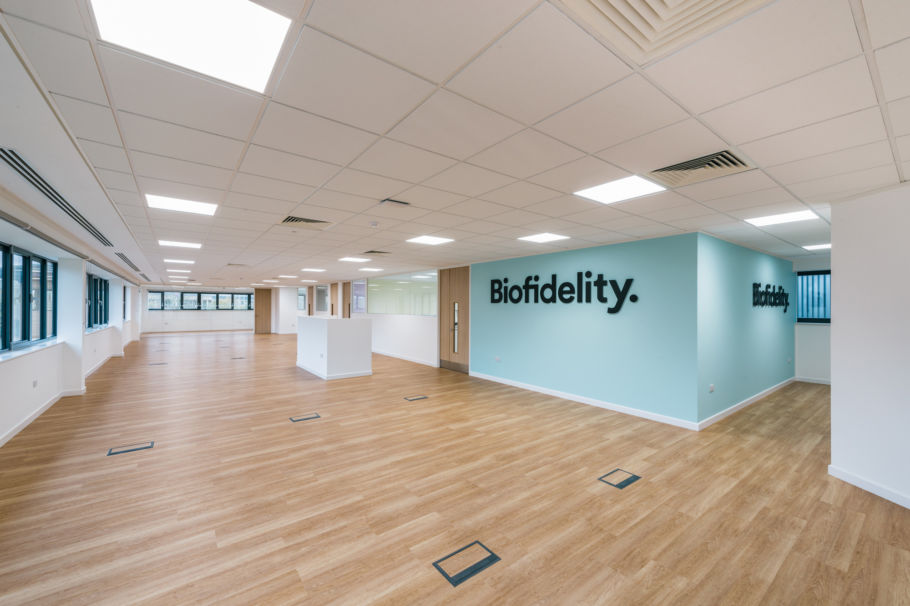 Spacious room with wooden flooring, white walls, and large windows features the text "Biofidelity" on a light blue wall, under a grid of ceiling lights.