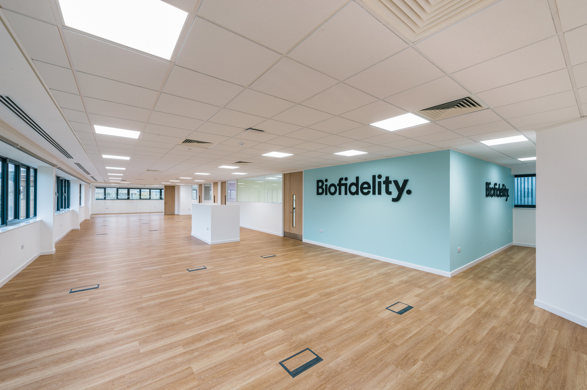 Spacious room with wooden flooring, white walls, and large windows features the text "Biofidelity" on a light blue wall, under a grid of ceiling lights.