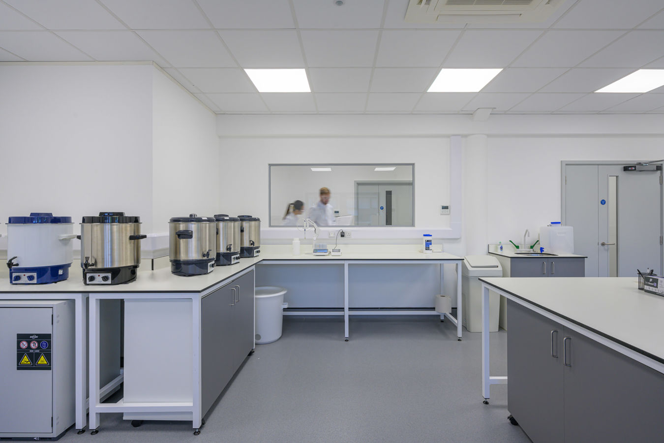 Laboratory equipment rests on countertops, including multiple large metallic containers; two people wearing lab coats can be seen through a window, suggesting a research environment in a bright, sterile room.