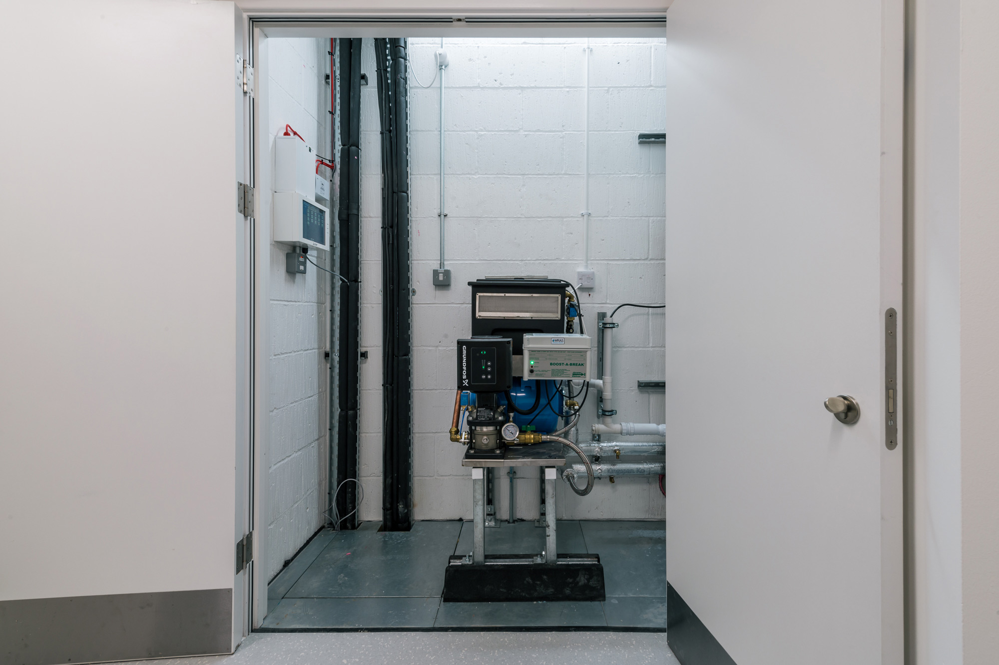 A pump system stands idle in a small, white-walled utility room with open doors, featuring visible pipes and electrical connections.