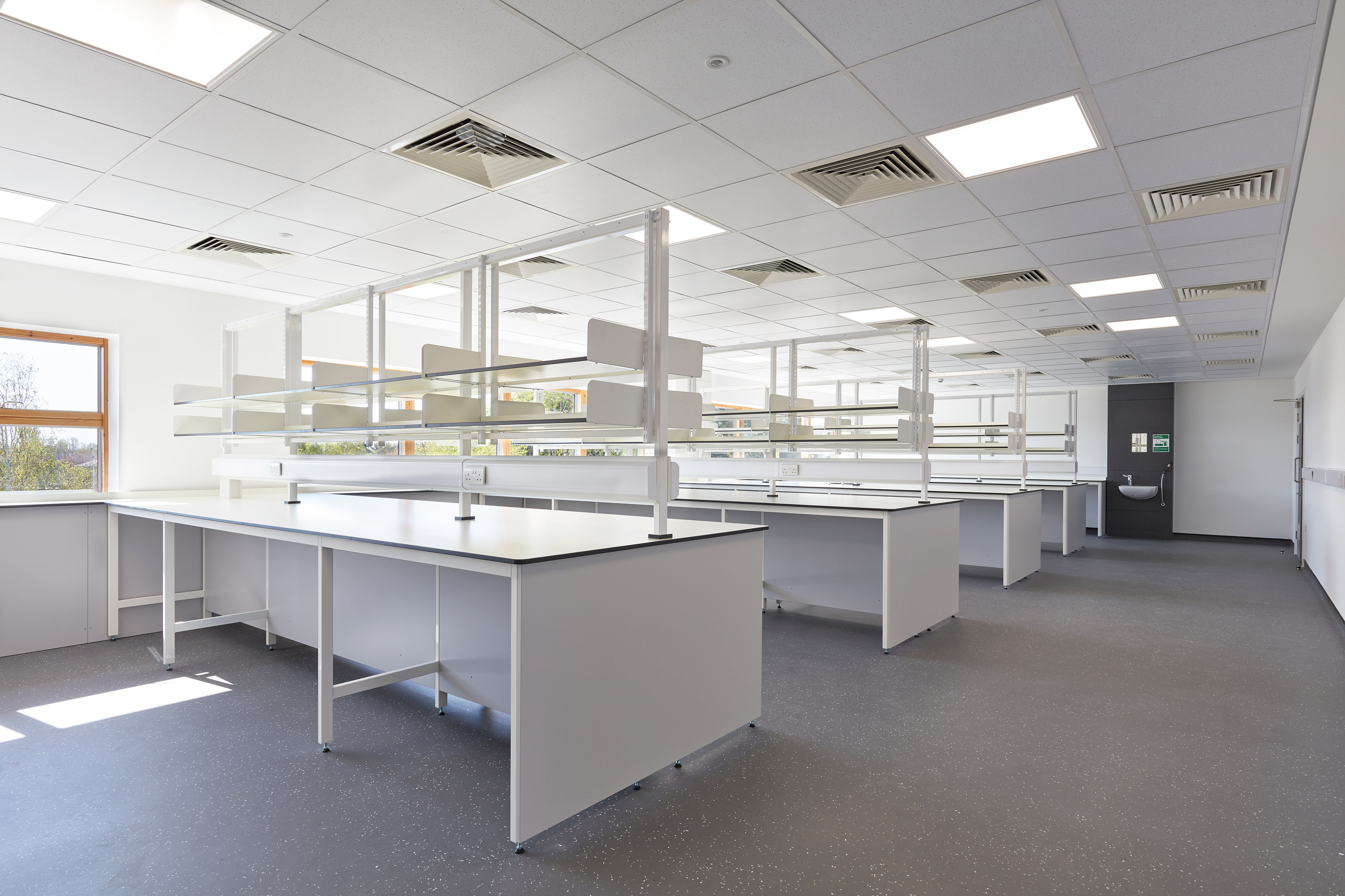 Cleanroom corridor with white walls and bright lighting, featuring lab personnel in protective clothing, wall-mounted lab coats, and a stainless steel bench and storage area.