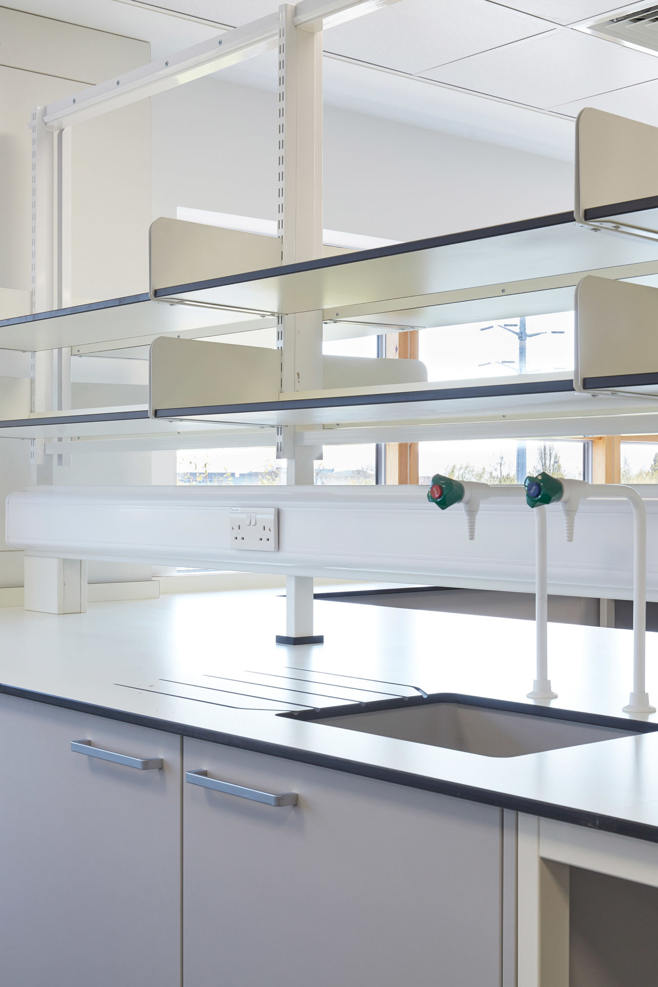 Bright and modern laboratory workspace featuring clean countertops, built-in cabinetry, glass shelving, and integrated water taps