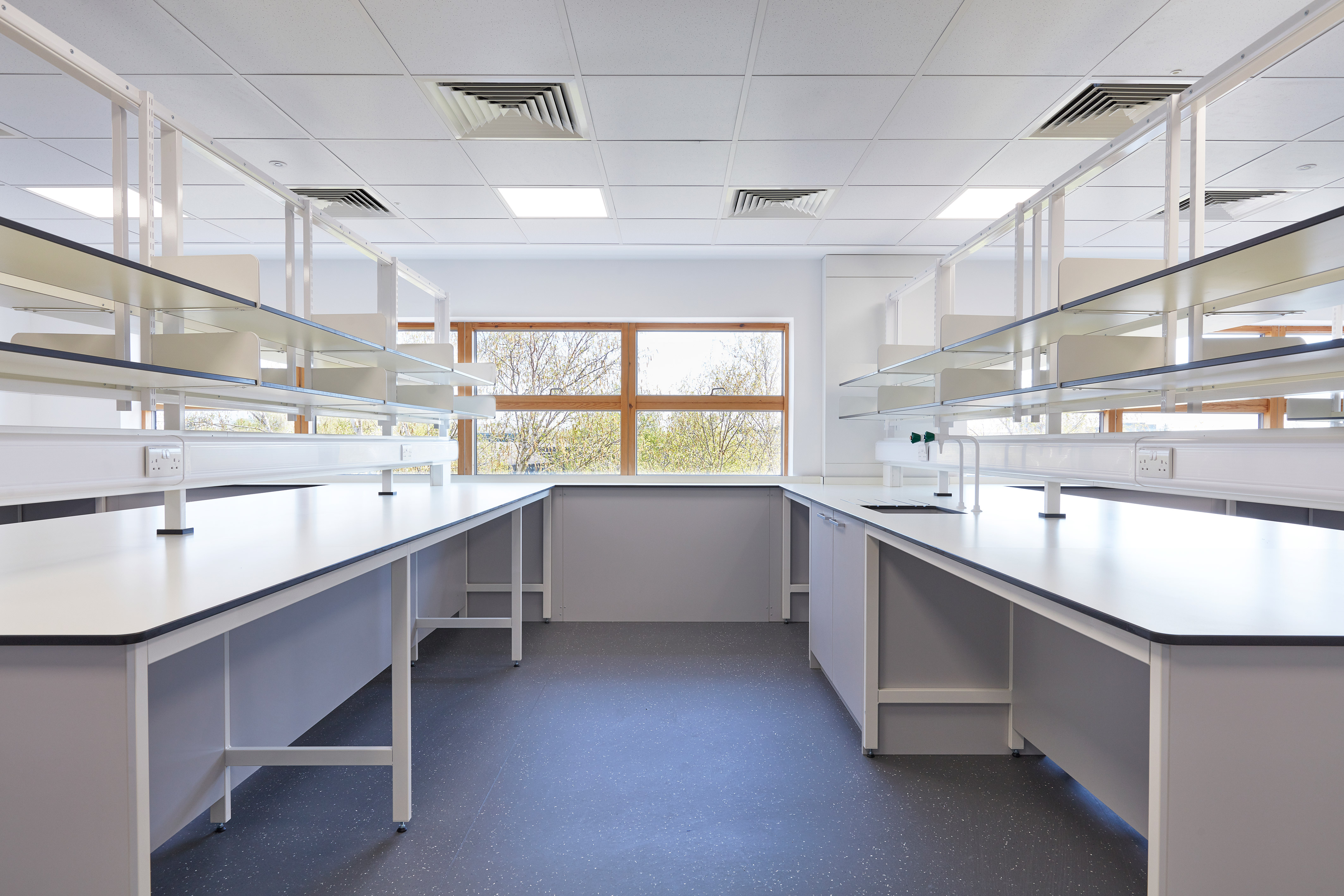 Bright laboratory with white workbenches, overhead shelving, blue flooring, and a large window providing natural light and outdoor views.