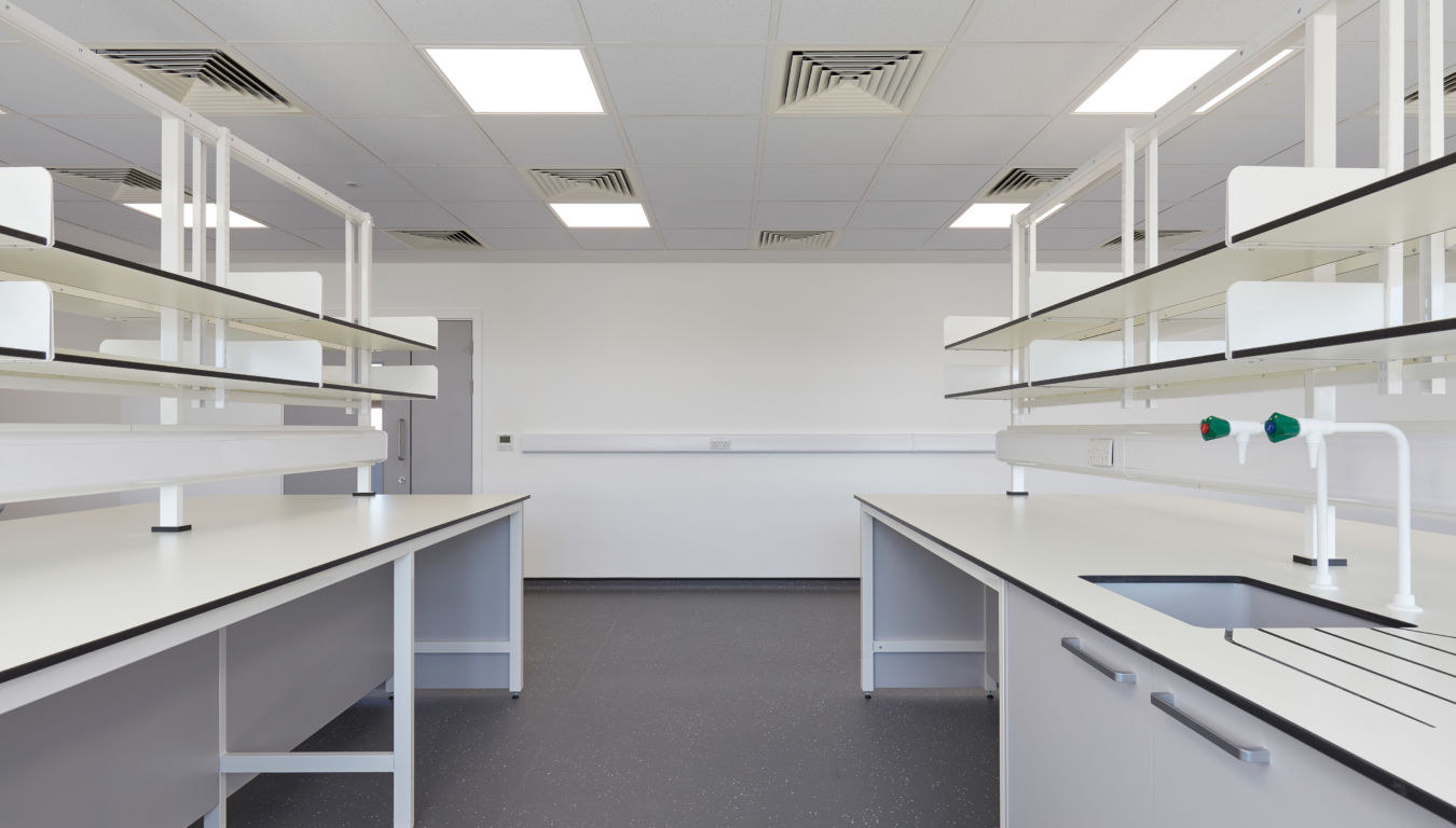 Modern laboratory interior with parallel white workbenches, overhead shelving units, bright ceiling lights, and dark flooring.