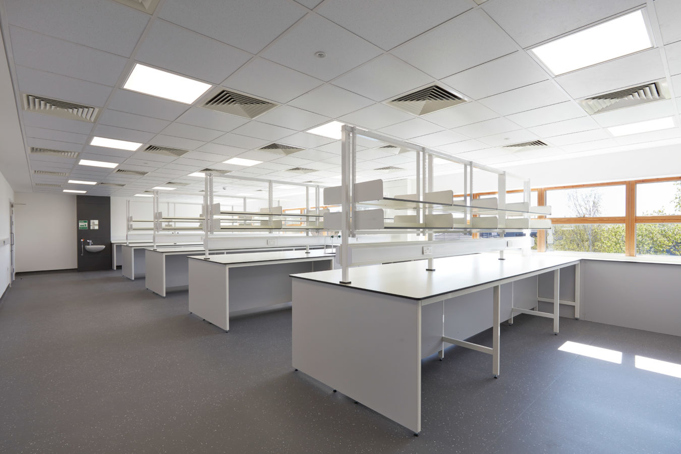 Spacious, well-lit laboratory with multiple white workbenches, overhead shelving units, and a grid of fluorescent ceiling lights.