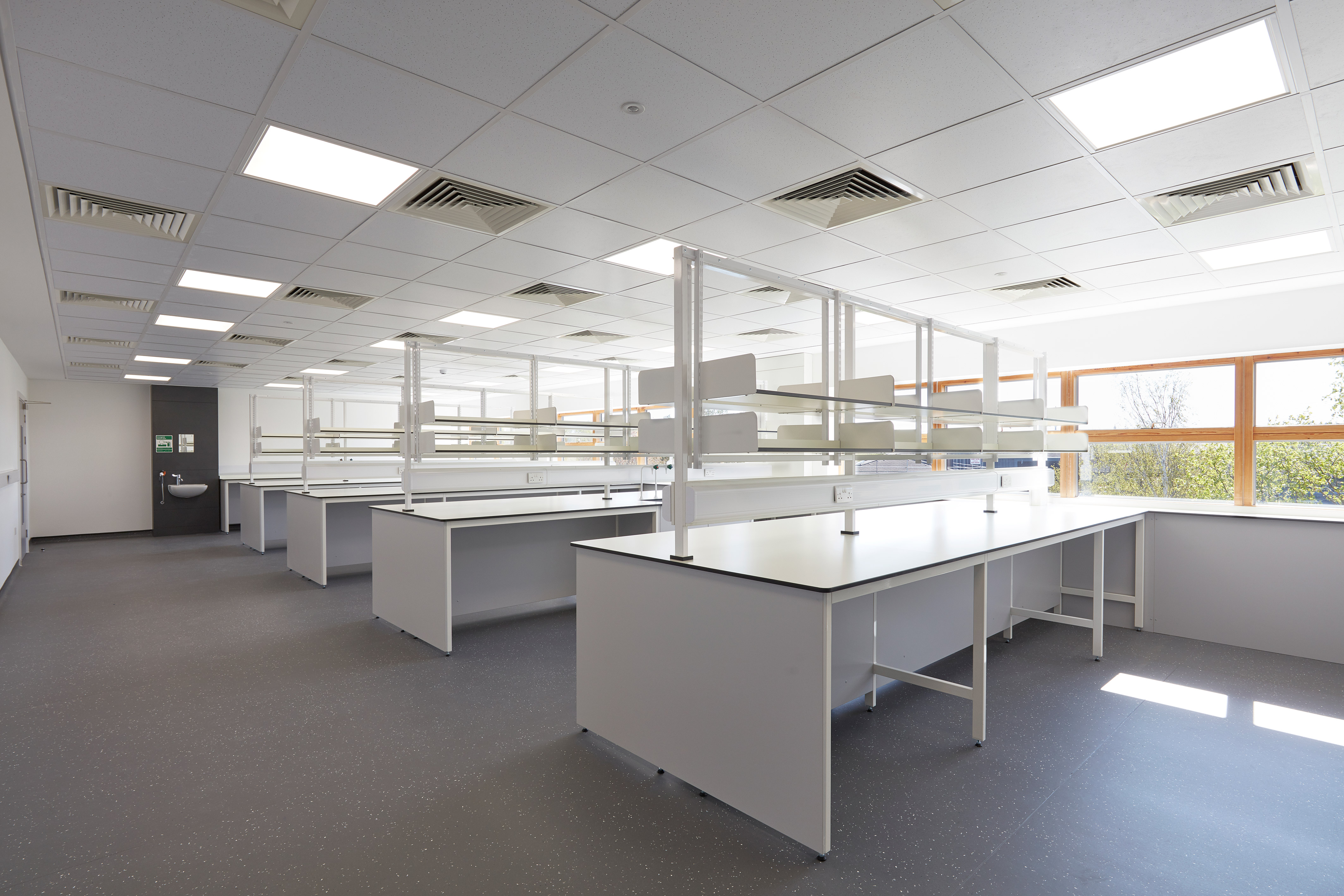 Spacious, well-lit laboratory with multiple white workbenches, overhead shelving units, and a grid of fluorescent ceiling lights.