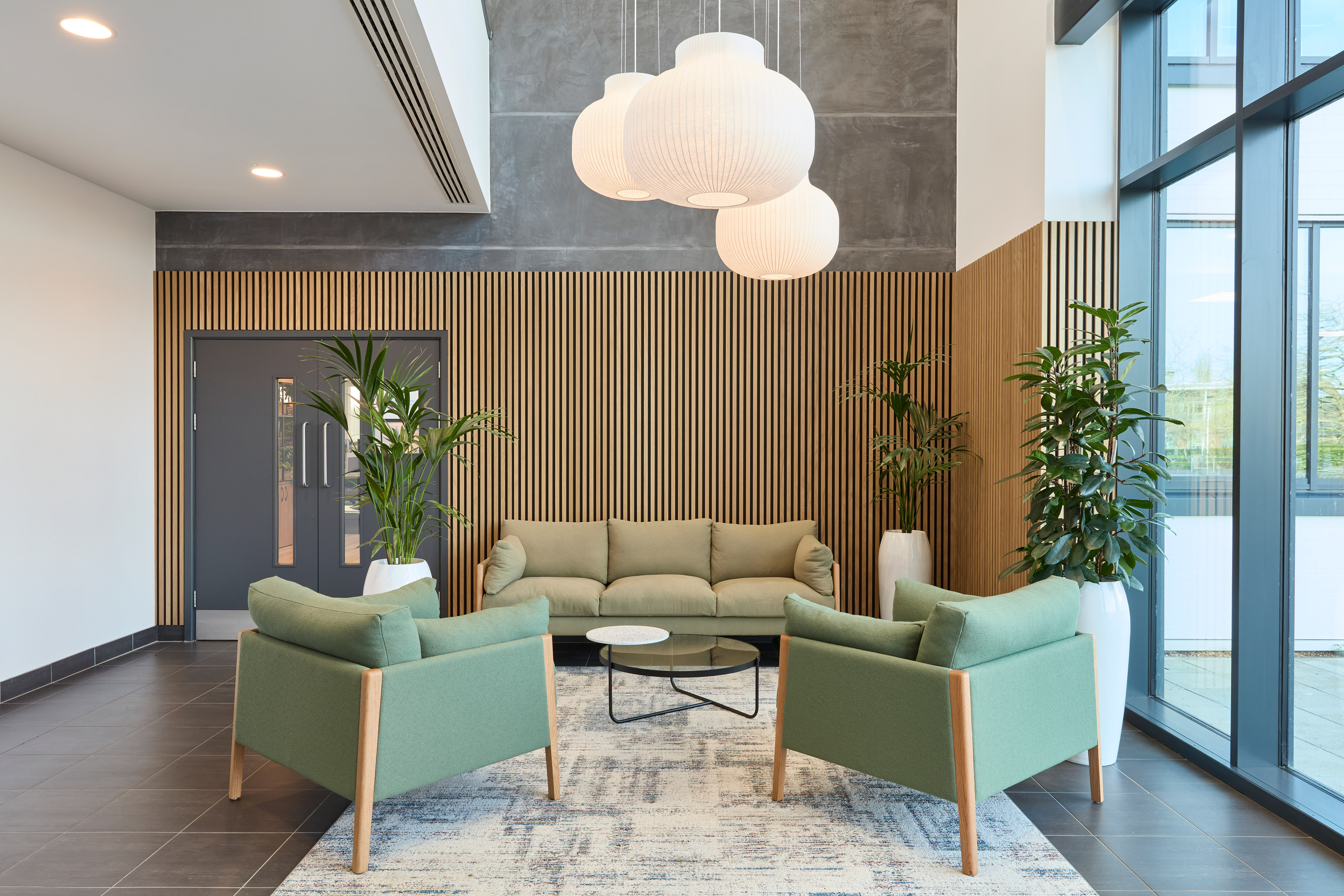 A modern lobby features green armchairs and a sofa around a glass coffee table, with large plants and pendant lights, surrounded by wooden paneling and large windows.