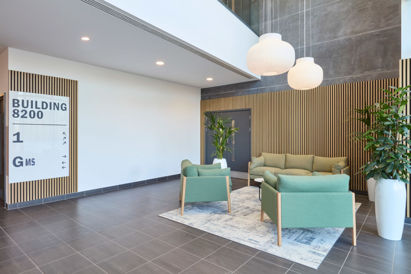 Seating area with green chairs and sofa on a rug; white pendant lights above. Sign reads "BUILDING 8200" with directional arrows. Potted plants against wood-paneled wall in an office lobby.
