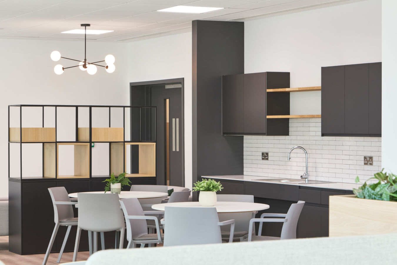 Modern kitchen area features a round table with chairs, contemporary open shelving, a sleek sink, and overhead cabinets against a tiled backsplash; plants accentuate the decor.