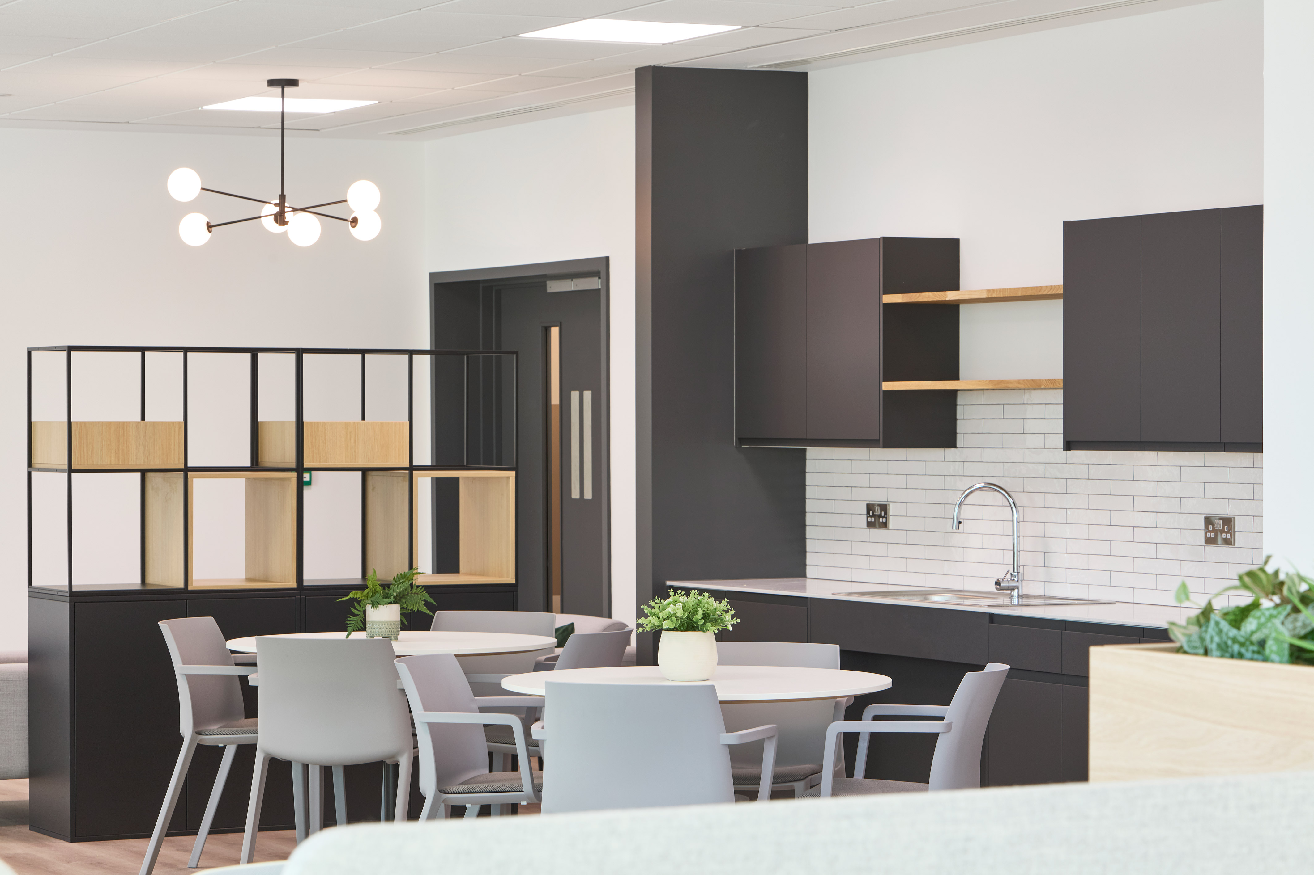 Modern kitchen area features a round table with chairs, contemporary open shelving, a sleek sink, and overhead cabinets against a tiled backsplash; plants accentuate the decor.
