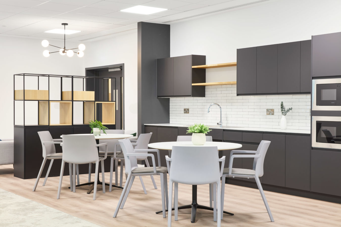 Modern kitchen area with gray cabinets and light countertops featuring a round table and chairs, decorative plants, and a tiled backsplash, illuminated by a minimalist overhead light fixture.