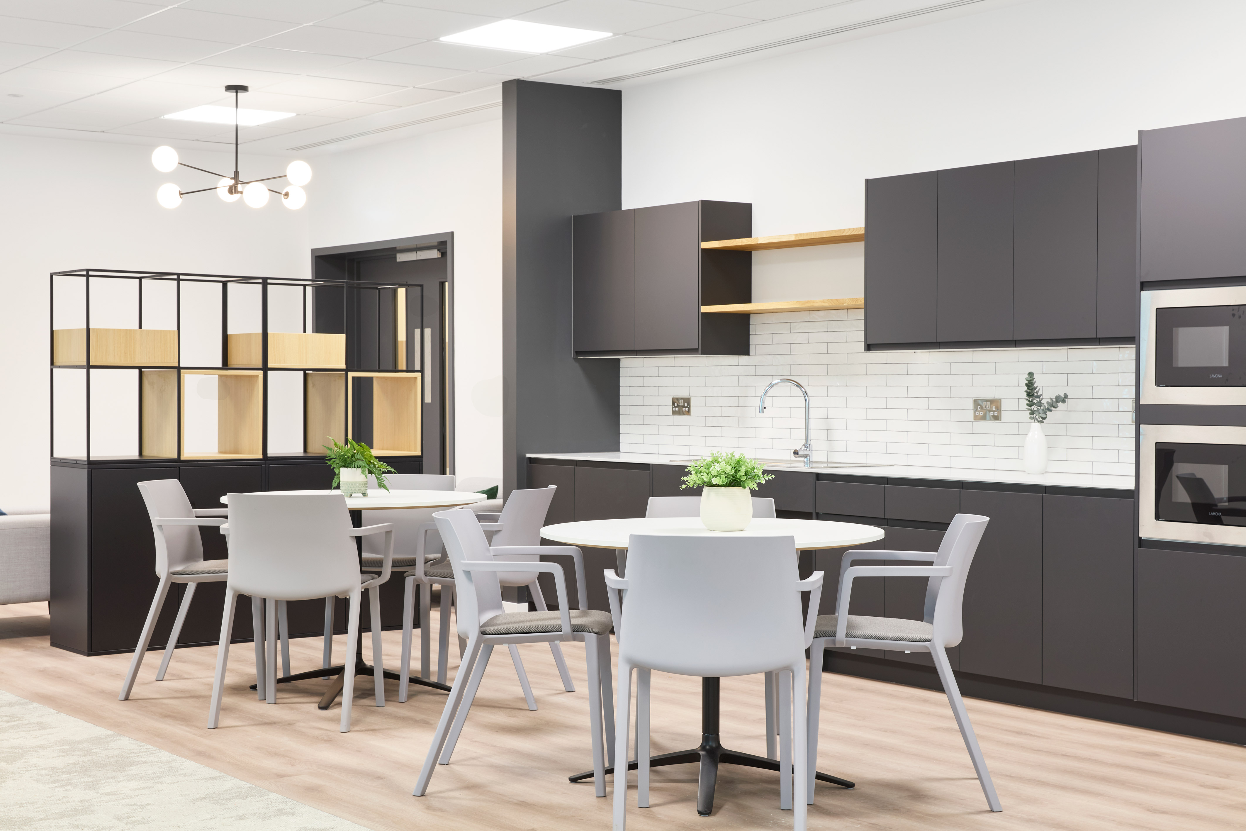 Modern kitchen area with gray cabinets and light countertops featuring a round table and chairs, decorative plants, and a tiled backsplash, illuminated by a minimalist overhead light fixture.