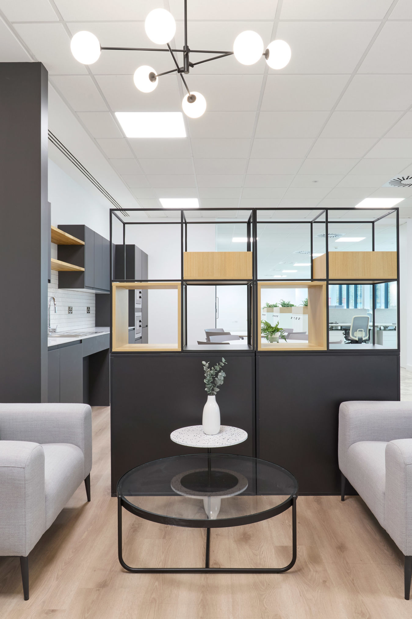 Modern office lounge with two gray sofas facing each other, accented by green and blue pillows. A black grid partition separates the space.