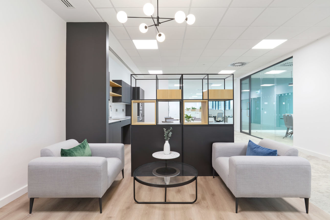 Modern office lounge with two gray sofas facing each other, accented by green and blue pillows. A black grid partition separates the space.