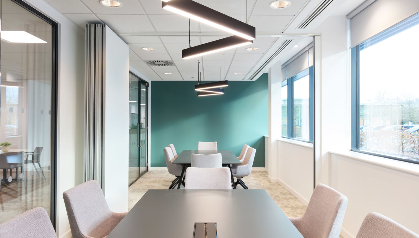 Modern conference room with light gray chairs around a black table, illuminated by sleek, angular pendant lights. The room has large windows and a teal accent wall.