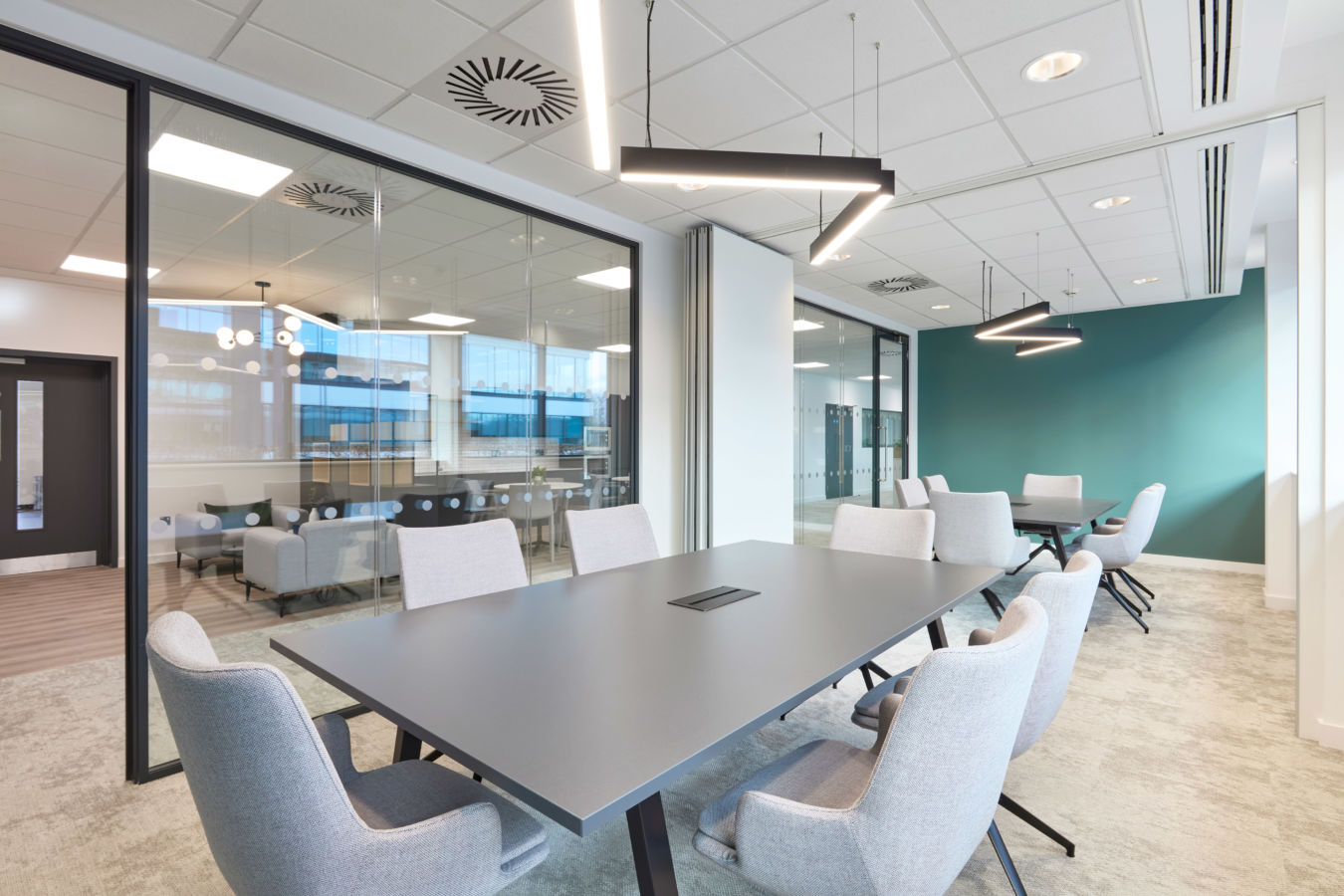 A modern conference room features a large gray table with six upholstered chairs, illuminated by sleek overhead lighting, set within a glass-walled office space with teal accent wall.