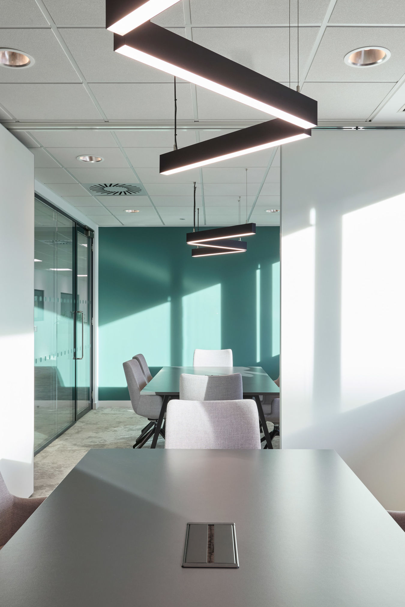 Ceiling lights illuminate a modern conference room with a long table, gray chairs, and a teal accent wall. Glass doors and sunlight create an airy, contemporary atmosphere.