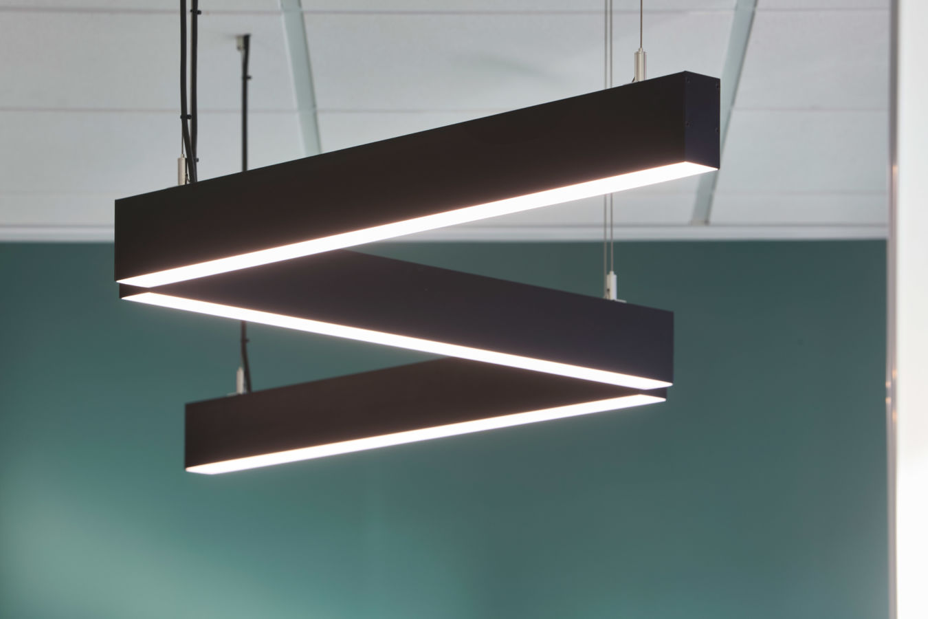 Suspended rectangular lighting fixture emits bright light, arranged in a zigzag pattern, with a clean, modern design against a teal wall in an office or commercial space.