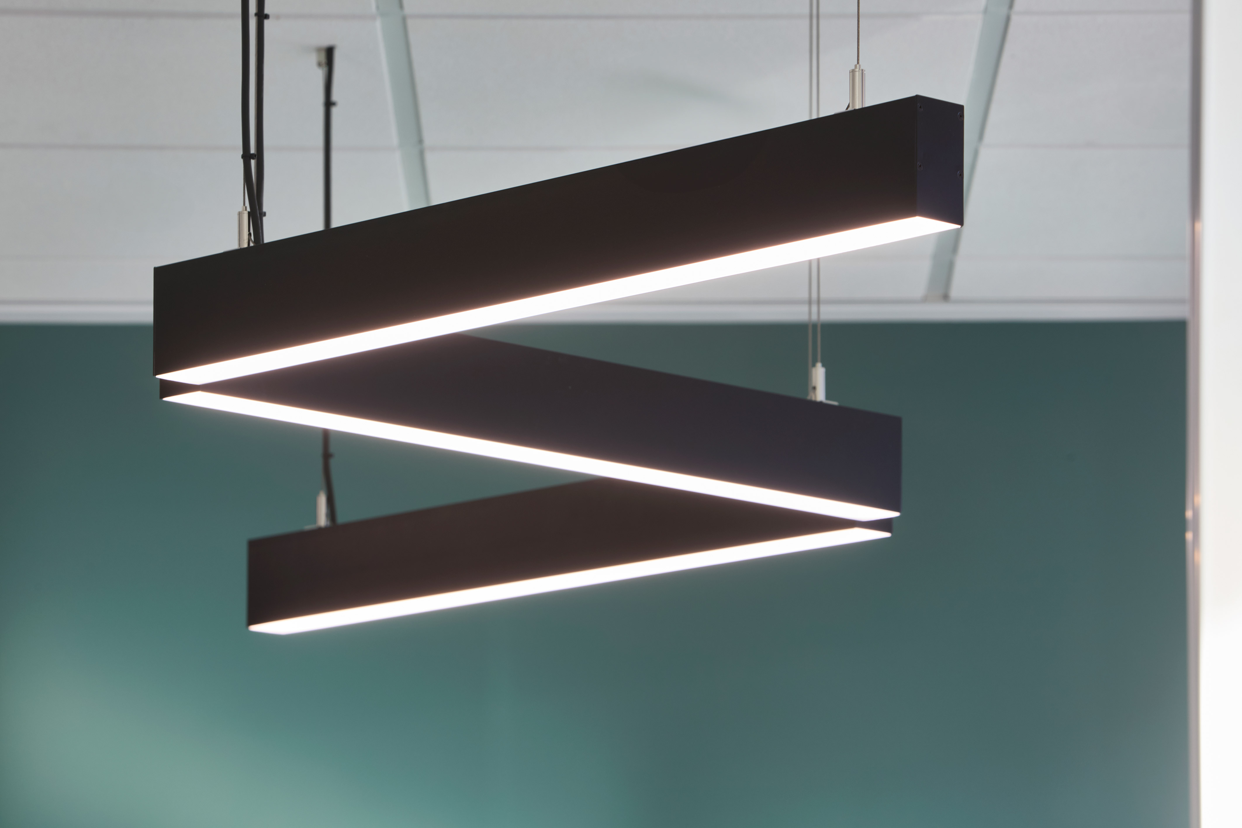 Suspended rectangular lighting fixture emits bright light, arranged in a zigzag pattern, with a clean, modern design against a teal wall in an office or commercial space.