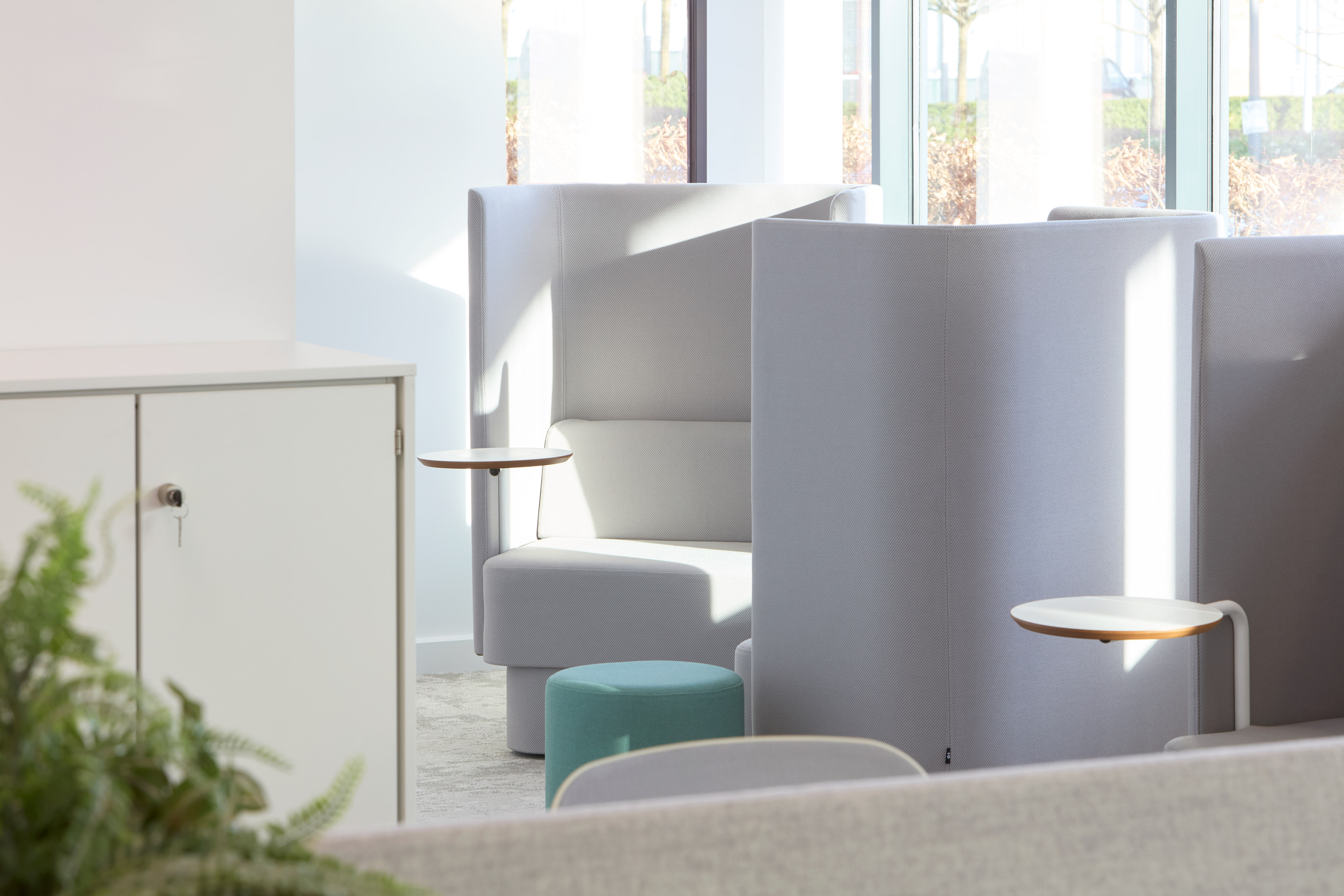 Gray high-backed chairs with integrated round side tables are arranged in a sunlit modern office space, complemented by a turquoise ottoman and a white cabinet with a visible key.