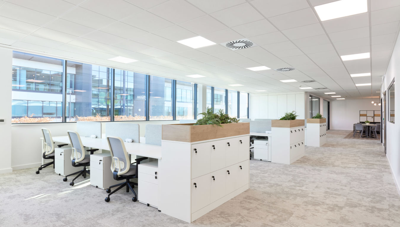 Spacious modern office with white desks, ergonomic chairs, and planters under bright lighting. Large windows offer an airy, organized ambiance