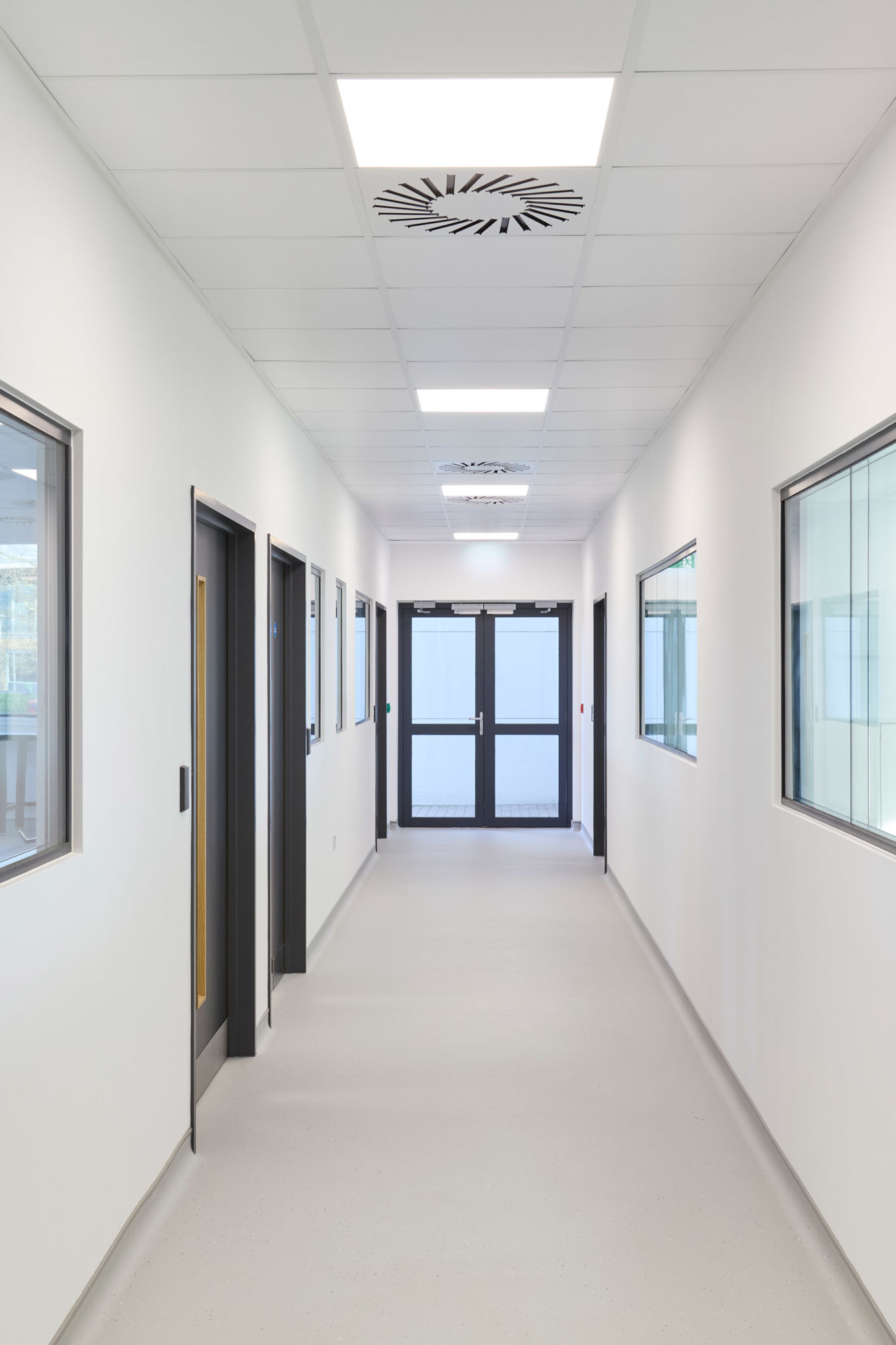 A clean, white corridor extends with closed black doors and windows lining the walls, under fluorescent ceiling lights, leading to double glass doors at the end.