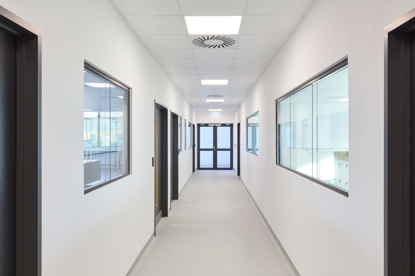 A clean, white corridor extends with closed black doors and windows lining the walls, under fluorescent ceiling lights, leading to double glass doors at the end.